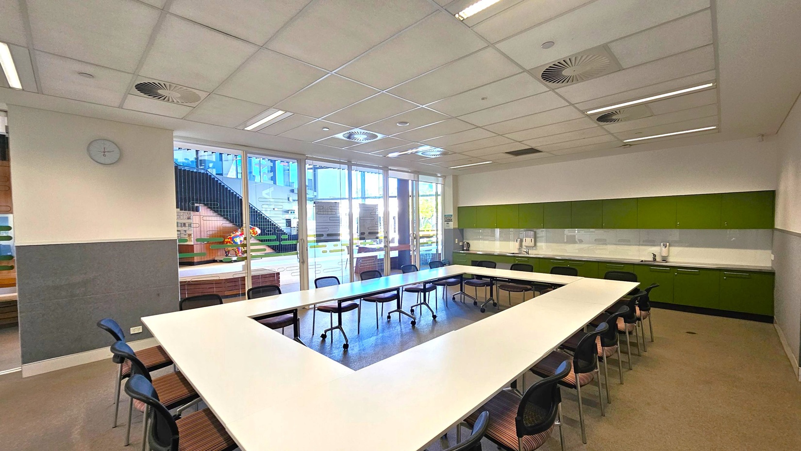 A room with a U-shaped arrangement of white tables and black chairs on a grey carpeted floor. The ceiling is fitted with white panels and circular vents. On the right, there is a long counter with green cabinets and a sink. The back wall features large glass doors with a view of an outdoor area, including stairs and colourful decorations. A clock is mounted on the left wall. A room with a U-shaped arrangement of white tables and black chairs on a grey carpeted floor. The ceiling is fitted with white panels and circular vents. On the right, there is a long counter with green cabinets and a sink. The back wall features large glass doors with a view of an outdoor area, including stairs and colourful decorations. A clock is mounted on the left wall.
