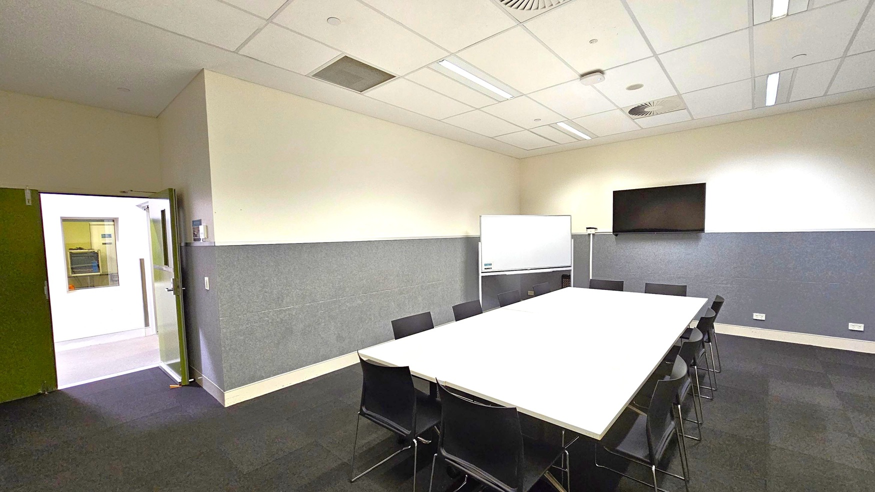 A conference room with a large rectangular table in the centre surrounded by black chairs. On the right wall, a flat-screen TV is mounted above a whiteboard. The walls are half grey and half cream, with a door open on the left leading to another room. The ceiling is fitted with fluorescent lights and ventilation panels. The floor is carpeted in dark grey tiles. A conference room with a large rectangular table in the centre surrounded by black chairs. On the right wall, a flat-screen TV is mounted above a whiteboard. The walls are half grey and half cream, with a door open on the left leading to another room. The ceiling is fitted with fluorescent lights and ventilation panels. The floor is carpeted in dark grey tiles.