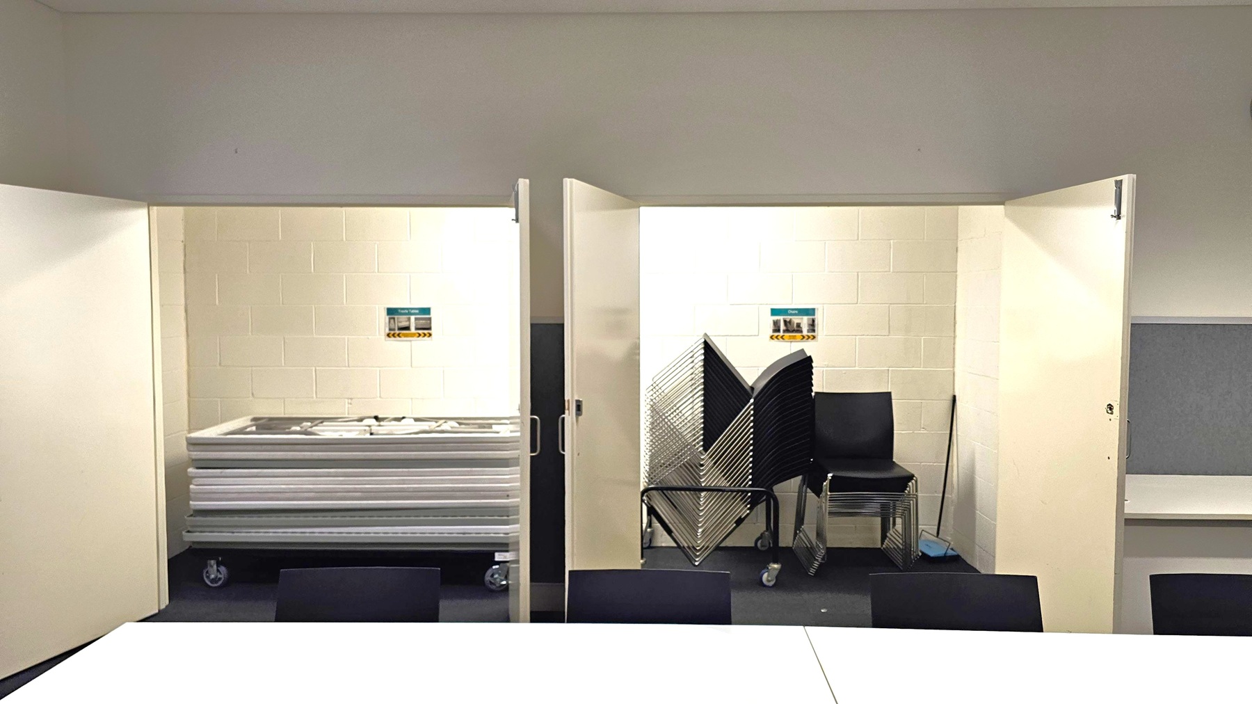 Two open storage areas with white walls. On the left, a stack of tables on wheels. On the right, a stack of chairs and folded metal racks. Both areas have small signs on the walls. The floor is dark, and the ceiling is light grey. In the foreground, a white table surface is partially visible. Two open storage areas with white walls. On the left, a stack of tables on wheels. On the right, a stack of chairs and folded metal racks. Both areas have small signs on the walls. The floor is dark, and the ceiling is light grey. In the foreground, a white table surface is partially visible.