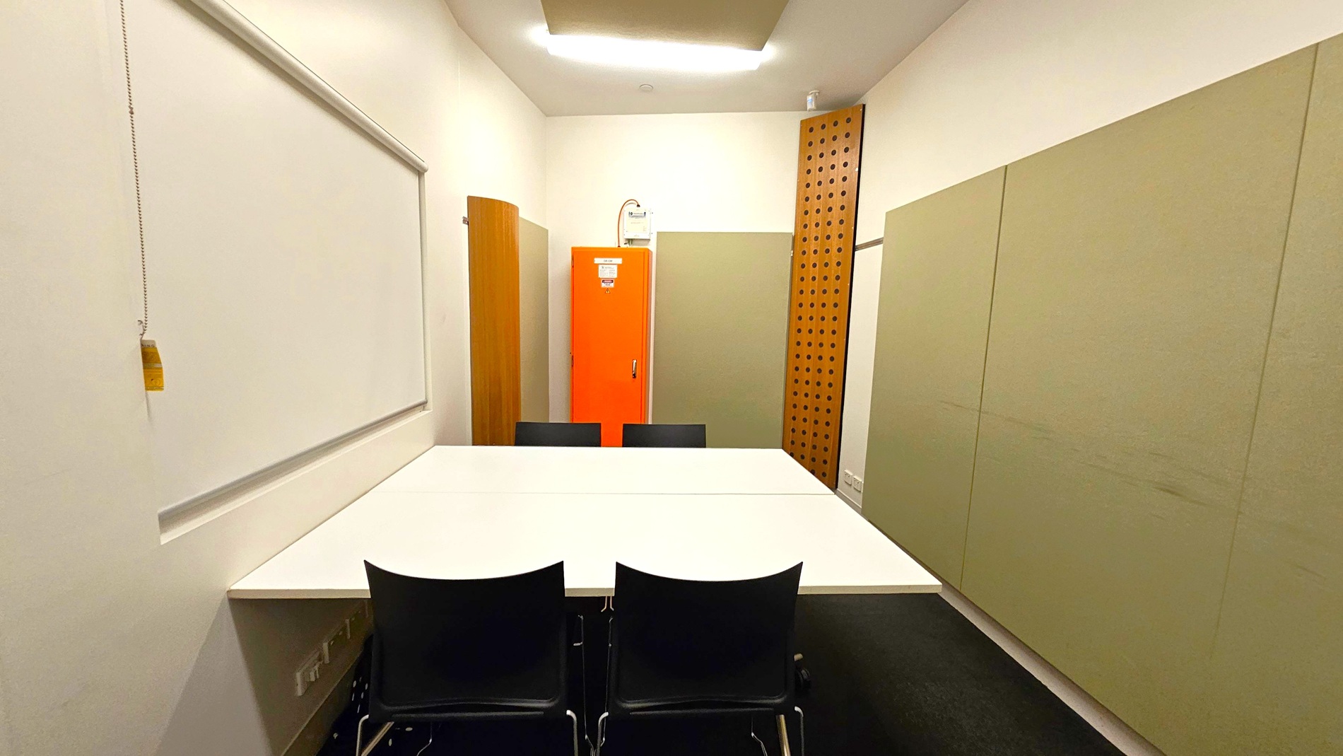 A small meeting room with a white table in the centre surrounded by four black chairs. On the left wall, there is a whiteboard with a pull chain. The back wall features an orange cabinet, flanked by beige panels and a wooden panel with circular cutouts. The floor is dark, and the ceiling has a rectangular light fixture. A small meeting room with a white table in the centre surrounded by four black chairs. On the left wall, there is a whiteboard with a pull chain. The back wall features an orange cabinet, flanked by beige panels and a wooden panel with circular cutouts. The floor is dark, and the ceiling has a rectangular light fixture.
