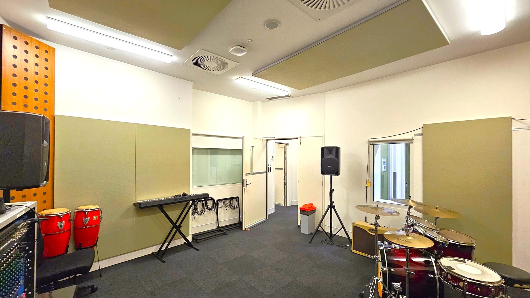 A music rehearsal room with a drum set on the right and a keyboard on a stand in the centre. On the left, there are red conga drums and a speaker. The walls are lined with beige acoustic panels, and there is a window with a blind partially open. The ceiling has fluorescent lights and ventilation panels. A door is open, leading to another room, and a waste bin is nearby. The floor is covered with dark carpet tiles. A music rehearsal room with a drum set on the right and a keyboard on a stand in the centre. On the left, there are red conga drums and a speaker. The walls are lined with beige acoustic panels, and there is a window with a blind partially open. The ceiling has fluorescent lights and ventilation panels. A door is open, leading to another room, and a waste bin is nearby. The floor is covered with dark carpet tiles.