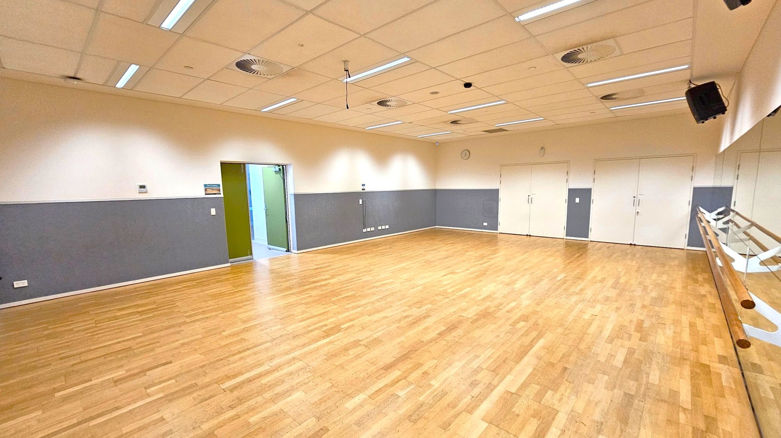 A spacious room with a wooden floor and a ceiling with recessed lighting and ventilation ducts. On the right, a mirrored wall with wooden ballet barres. The left wall is grey with a doorway leading to another area. The back wall has several white doors and a clock. The room is well-lit and appears to be a dance or exercise studio. A spacious room with a wooden floor and a ceiling with recessed lighting and ventilation ducts. On the right, a mirrored wall with wooden ballet barres. The left wall is grey with a doorway leading to another area. The back wall has several white doors and a clock. The room is well-lit and appears to be a dance or exercise studio.