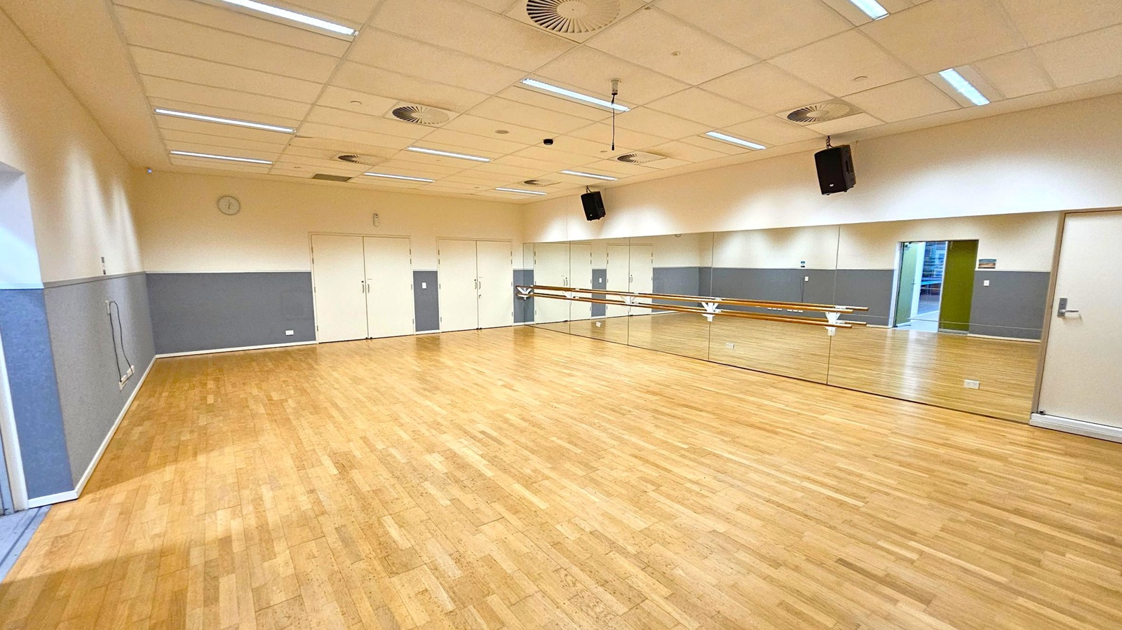 A spacious dance studio with a wooden floor and a mirrored wall on the right, featuring a ballet barre. The left wall is painted grey and white, with a few electrical outlets visible. The back wall has several closed doors. The ceiling is fitted with fluorescent lights and air vents. A speaker is mounted on the right wall. An open doorway on the right leads to another area. A spacious dance studio with a wooden floor and a mirrored wall on the right, featuring a ballet barre. The left wall is painted grey and white, with a few electrical outlets visible. The back wall has several closed doors. The ceiling is fitted with fluorescent lights and air vents. A speaker is mounted on the right wall. An open doorway on the right leads to another area.