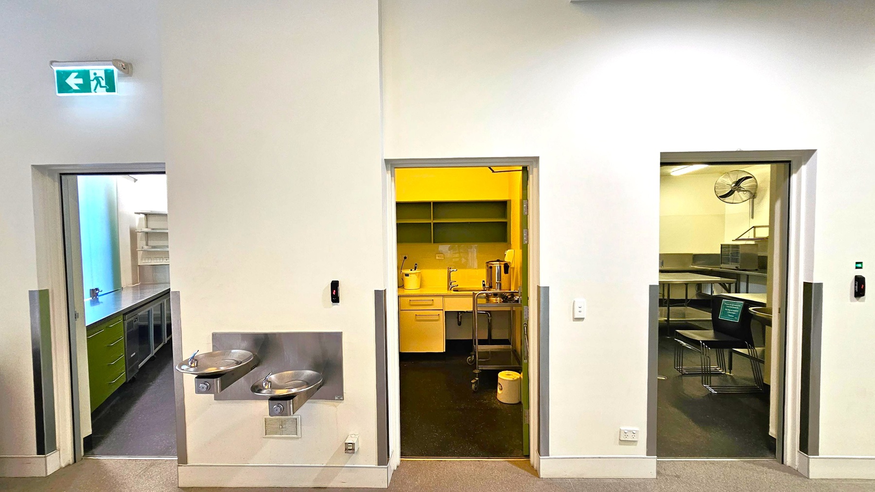 A wall with three doorways. The left doorway leads to a room with shelves and a countertop. Above it, an illuminated green exit sign with a left arrow. In the centre, a doorway opens to a kitchenette with a sink, shelves, and a kettle. Below this doorway, two stainless steel drinking fountains are mounted on the wall. The right doorway reveals a room with tables, chairs, and a wall-mounted fan. The floor is carpeted, and the walls are light-coloured. A wall with three doorways. The left doorway leads to a room with shelves and a countertop. Above it, an illuminated green exit sign with a left arrow. In the centre, a doorway opens to a kitchenette with a sink, shelves, and a kettle. Below this doorway, two stainless steel drinking fountains are mounted on the wall. The right doorway reveals a room with tables, chairs, and a wall-mounted fan. The floor is carpeted, and the walls are light-coloured.