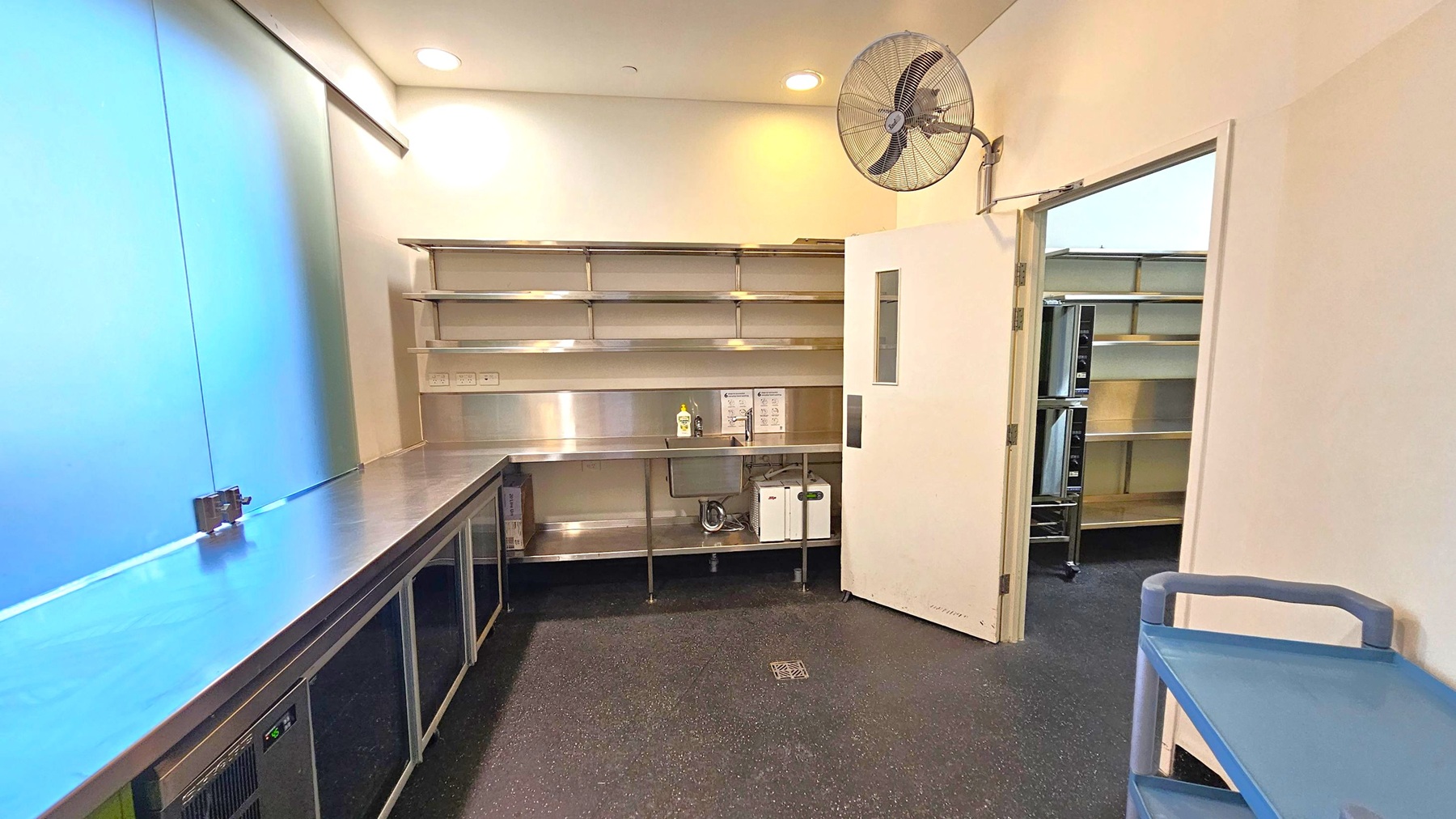 A commercial kitchen with stainless steel countertops and shelving on the left. Below the counter are refrigeration units. On the right, a door is partially open, revealing more shelving in the adjacent room. A wall-mounted fan is above the door. The floor is dark with a speckled pattern, and the ceiling is white with recessed lighting. A blue trolley is in the foreground on the right. A commercial kitchen with stainless steel countertops and shelving on the left. Below the counter are refrigeration units. On the right, a door is partially open, revealing more shelving in the adjacent room. A wall-mounted fan is above the door. The floor is dark with a speckled pattern, and the ceiling is white with recessed lighting. A blue trolley is in the foreground on the right.