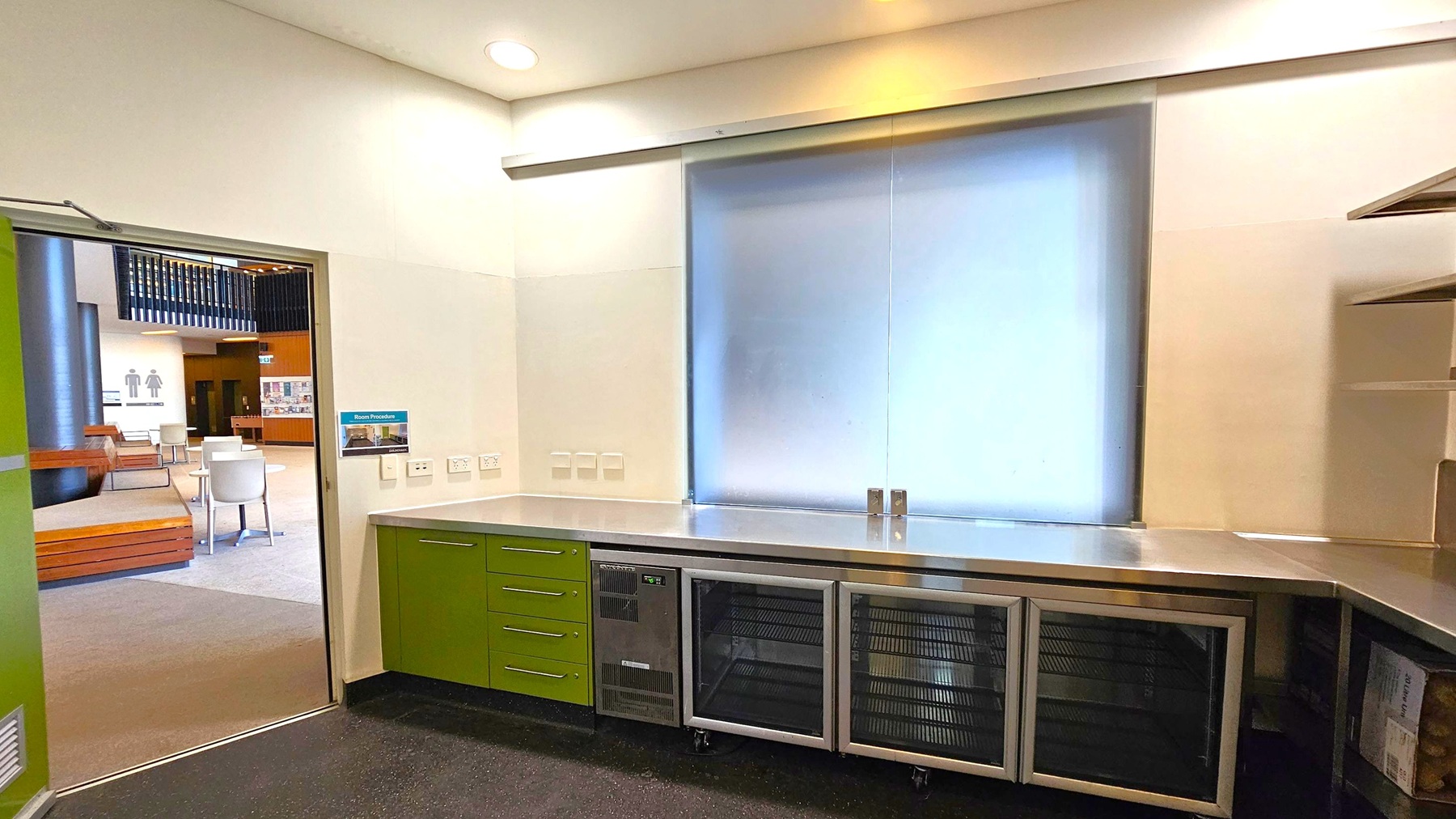 A room with a stainless steel countertop and green cabinets below. There are multiple power outlets on the wall above the counter. A large frosted window is centred on the wall. To the left, an open door leads to a lobby area with chairs and tables. Signs for restrooms are visible in the background. The floor is dark and speckled, and the ceiling is light with recessed lighting. Shelving is partially visible on the right. A room with a stainless steel countertop and green cabinets below. There are multiple power outlets on the wall above the counter. A large frosted window is centred on the wall. To the left, an open door leads to a lobby area with chairs and tables. Signs for restrooms are visible in the background. The floor is dark and speckled, and the ceiling is light with recessed lighting. Shelving is partially visible on the right.