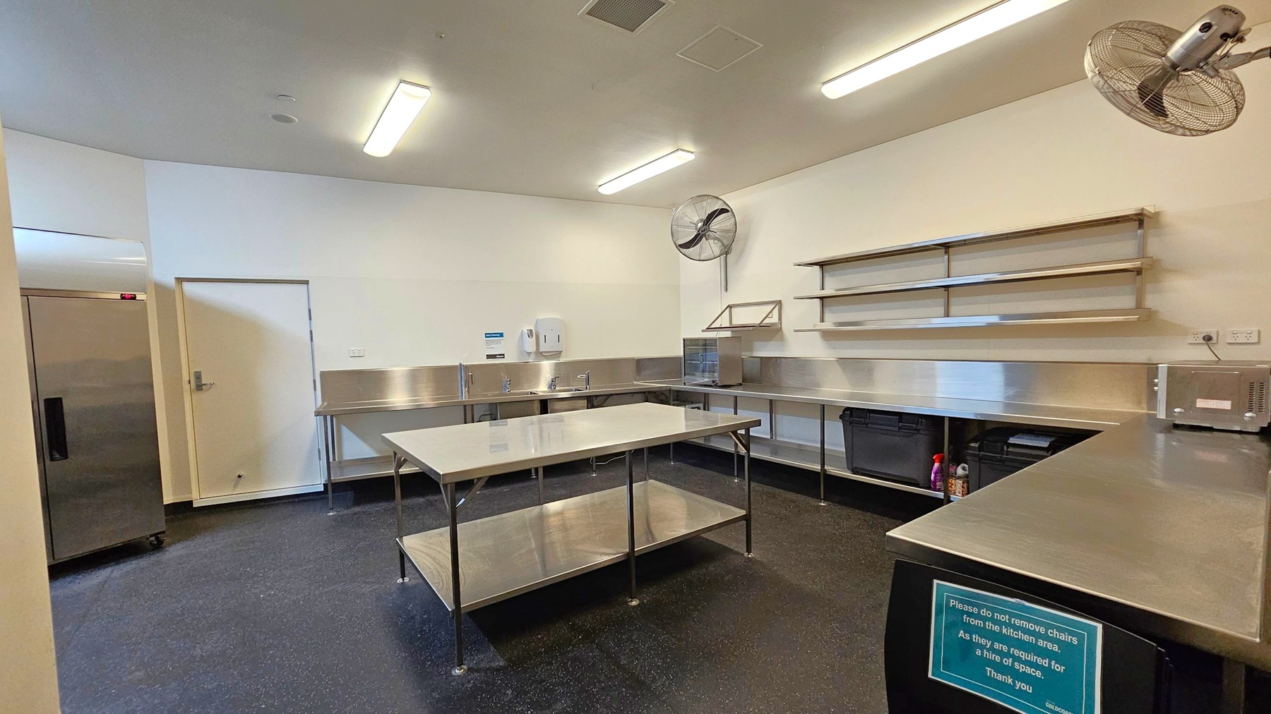 A commercial kitchen with stainless steel worktops and shelving on the right wall. In the centre, there is a large stainless steel table with a lower shelf. On the left, a closed door and a large refrigerator. The floor is dark and speckled, while the ceiling is white with fluorescent lights and wall-mounted fans. A sign on the front worktop advises not to remove chairs from the kitchen area. A commercial kitchen with stainless steel worktops and shelving on the right wall. In the centre, there is a large stainless steel table with a lower shelf. On the left, a closed door and a large refrigerator. The floor is dark and speckled, while the ceiling is white with fluorescent lights and wall-mounted fans. A sign on the front worktop advises not to remove chairs from the kitchen area.
