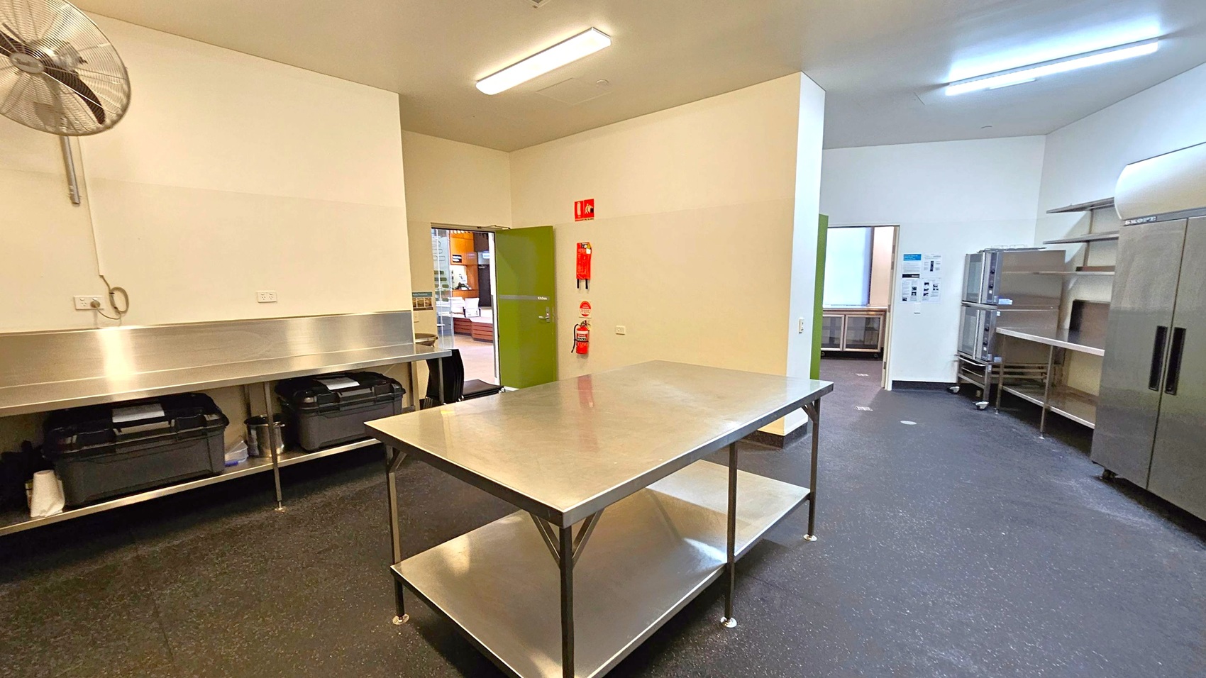 A commercial kitchen with a stainless steel worktable in the centre. On the left, a wall-mounted fan and a long counter with storage underneath. In the background, a green door leads to another room, with fire safety equipment mounted nearby. On the right, stainless steel shelves and a large refrigerator. The floor is dark and speckled, and the ceiling has fluorescent lighting. A commercial kitchen with a stainless steel worktable in the centre. On the left, a wall-mounted fan and a long counter with storage underneath. In the background, a green door leads to another room, with fire safety equipment mounted nearby. On the right, stainless steel shelves and a large refrigerator. The floor is dark and speckled, and the ceiling has fluorescent lighting.