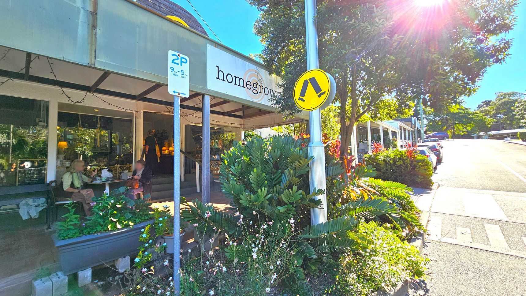 A café with a sign reading "homegrown" on the left, featuring outdoor seating where two people are sitting at a table. A parking sign and a pedestrian crossing sign are visible near lush green plants. On the right, a street lined with parked cars extends uphill, bordered by trees and more greenery. Sunlight filters through the trees, casting shadows on the pavement. A café with a sign reading "homegrown" on the left, featuring outdoor seating where two people are sitting at a table. A parking sign and a pedestrian crossing sign are visible near lush green plants. On the right, a street lined with parked cars extends uphill, bordered by trees and more greenery. Sunlight filters through the trees, casting shadows on the pavement.