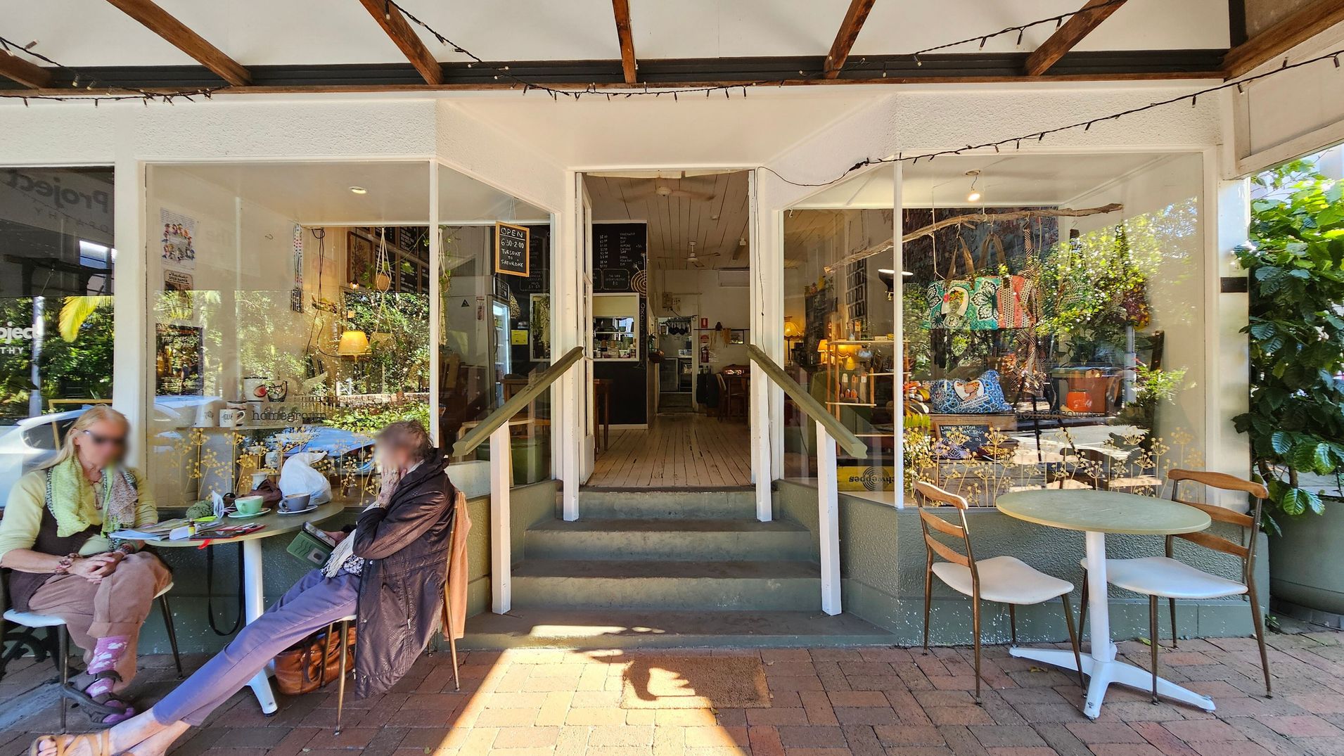 A small café entrance with two large windows on either side. On the left, two people sit at a round table with drinks and papers. On the right, another round table with two empty chairs. Inside, a wooden floor leads to a counter with a menu board above. The windows display colourful bags and plants. The ceiling has exposed wooden beams with string lights, and the floor is brick. A small café entrance with two large windows on either side. On the left, two people sit at a round table with drinks and papers. On the right, another round table with two empty chairs. Inside, a wooden floor leads to a counter with a menu board above. The windows display colourful bags and plants. The ceiling has exposed wooden beams with string lights, and the floor is brick.
