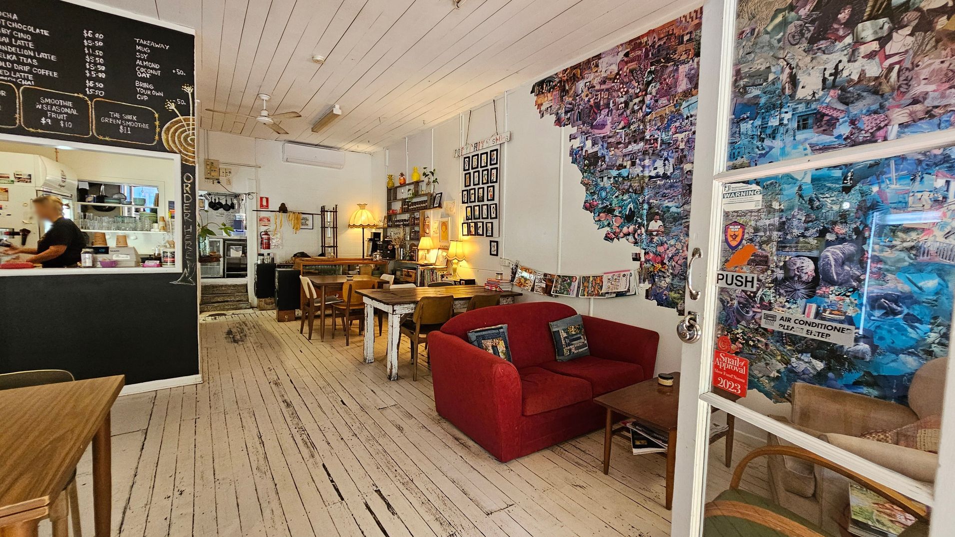 A cosy café interior with a rustic wooden floor and white panelled ceiling. On the left, a blackboard menu is displayed above a counter where a person is working. In the centre, wooden tables and chairs are arranged, with a vintage lamp and shelves in the background. On the right, a red sofa and a collage-covered wall add a colourful touch. A glass door with stickers and a "PUSH" sign is visible, leading to another seating area. A cosy café interior with a rustic wooden floor and white panelled ceiling. On the left, a blackboard menu is displayed above a counter where a person is working. In the centre, wooden tables and chairs are arranged, with a vintage lamp and shelves in the background. On the right, a red sofa and a collage-covered wall add a colourful touch. A glass door with stickers and a "PUSH" sign is visible, leading to another seating area.