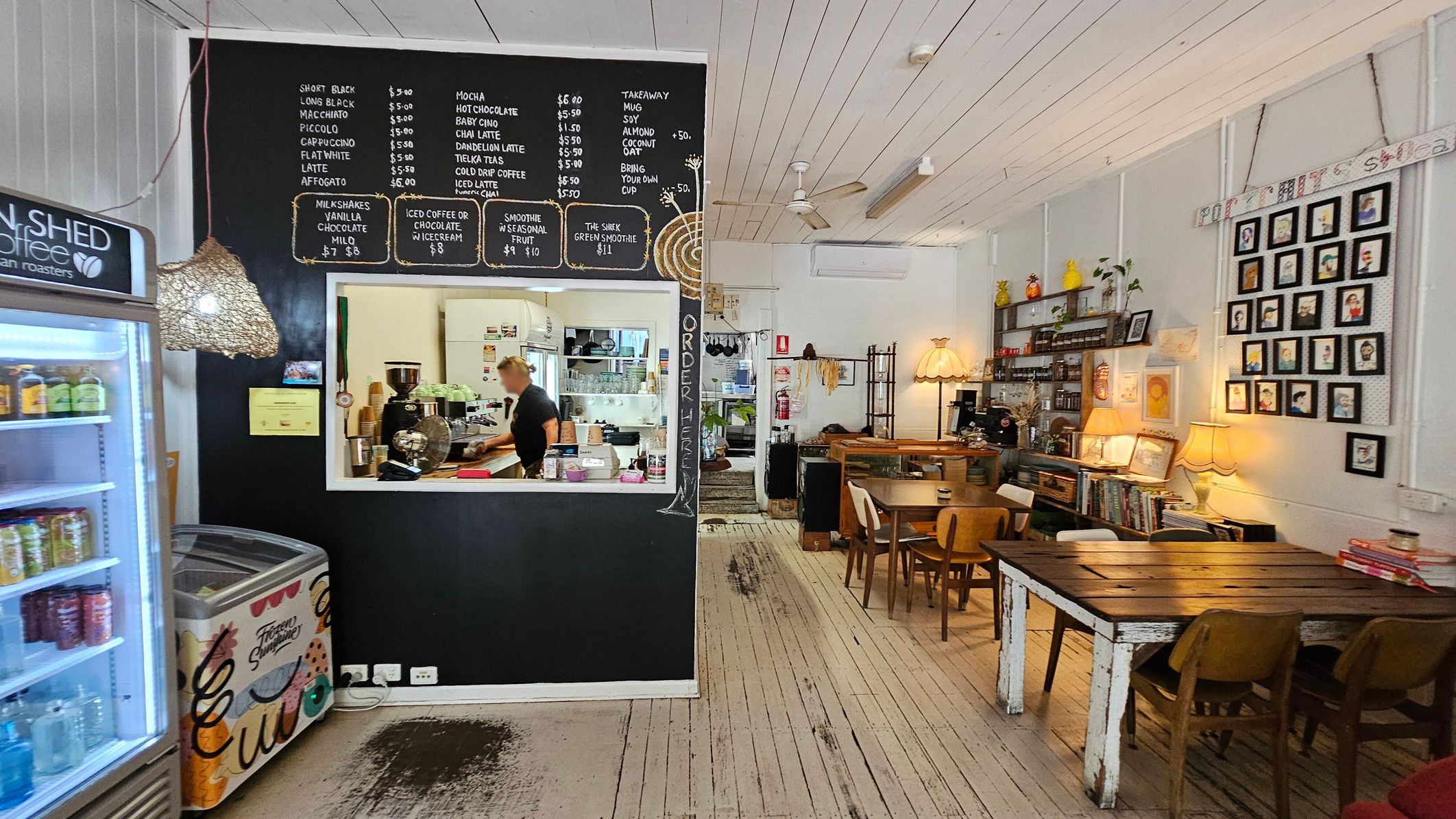 A cosy café interior with a blackboard menu on the left wall above a serving window, where a person is preparing drinks. A fridge with drinks and an ice cream freezer are nearby. On the right, wooden tables and chairs are arranged, with a wall displaying framed pictures. Shelves hold various items, and warm lighting from lamps creates a welcoming atmosphere. The floor is wooden, and the ceiling is white with exposed beams and a ceiling fan. A cosy café interior with a blackboard menu on the left wall above a serving window, where a person is preparing drinks. A fridge with drinks and an ice cream freezer are nearby. On the right, wooden tables and chairs are arranged, with a wall displaying framed pictures. Shelves hold various items, and warm lighting from lamps creates a welcoming atmosphere. The floor is wooden, and the ceiling is white with exposed beams and a ceiling fan.