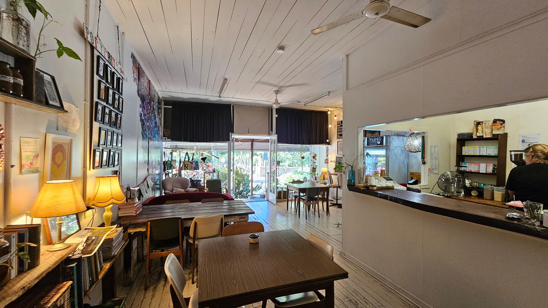 A cosy café interior with wooden flooring and a white panelled ceiling. On the left, a wall displays framed photos and artwork, with a shelf holding books and a lamp. In the centre, tables and chairs are arranged, leading to a glass entrance with views of greenery outside. On the right, a counter with a person working behind it, shelves stocked with products, and a hanging light fixture. Ceiling fans are visible above. A cosy café interior with wooden flooring and a white panelled ceiling. On the left, a wall displays framed photos and artwork, with a shelf holding books and a lamp. In the centre, tables and chairs are arranged, leading to a glass entrance with views of greenery outside. On the right, a counter with a person working behind it, shelves stocked with products, and a hanging light fixture. Ceiling fans are visible above.