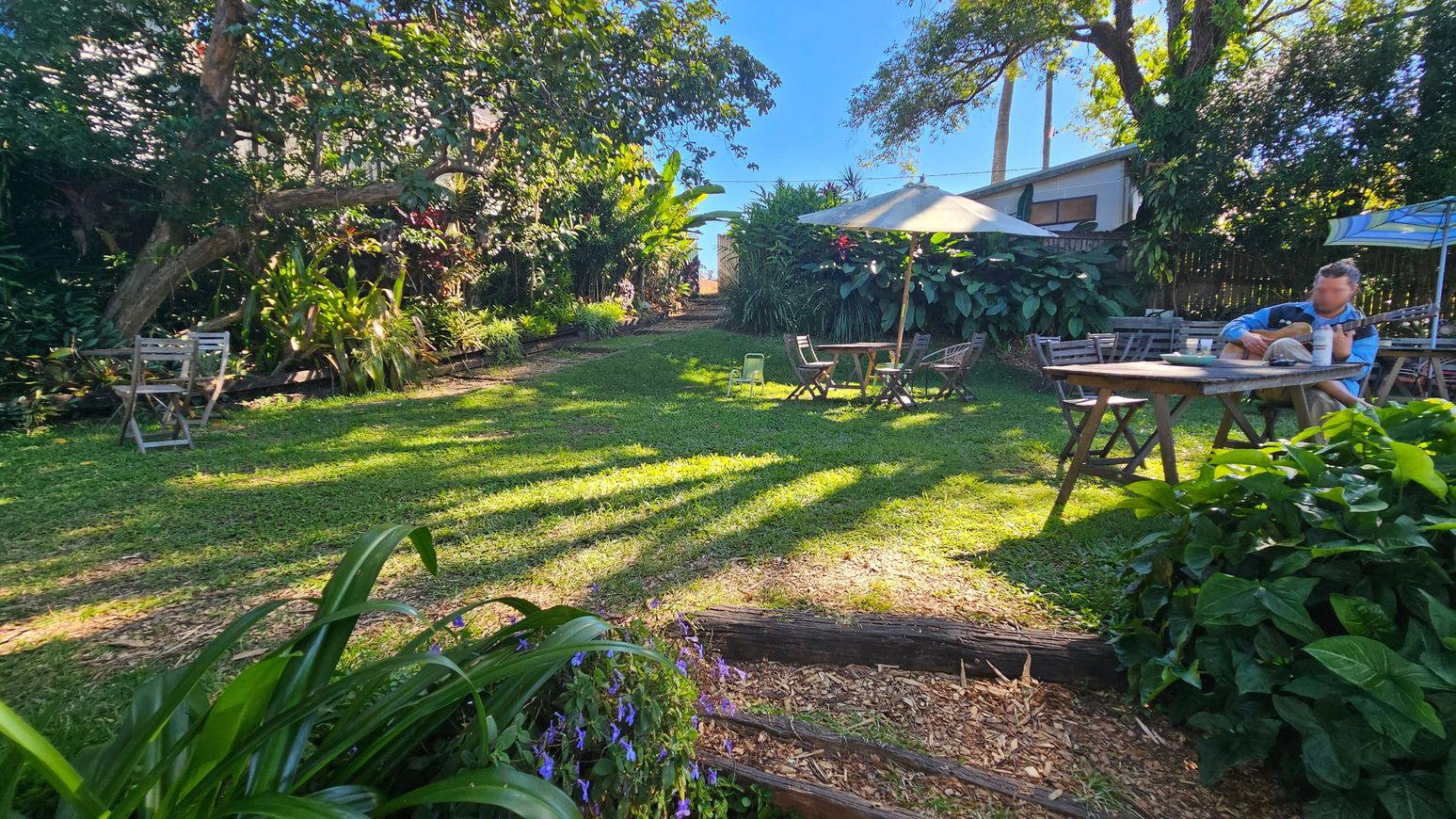 A garden with a grassy area surrounded by lush greenery and trees. On the left, there are two wooden chairs near dense foliage. In the centre, a pathway leads through the garden, with a table and chairs under a large umbrella. On the right, a person is seated at a wooden table playing a guitar. The foreground has vibrant plants and flowers, while the background shows more trees and a wooden fence. Shadows from the trees stretch across the grass. A garden with a grassy area surrounded by lush greenery and trees. On the left, there are two wooden chairs near dense foliage. In the centre, a pathway leads through the garden, with a table and chairs under a large umbrella. On the right, a person is seated at a wooden table playing a guitar. The foreground has vibrant plants and flowers, while the background shows more trees and a wooden fence. Shadows from the trees stretch across the grass.