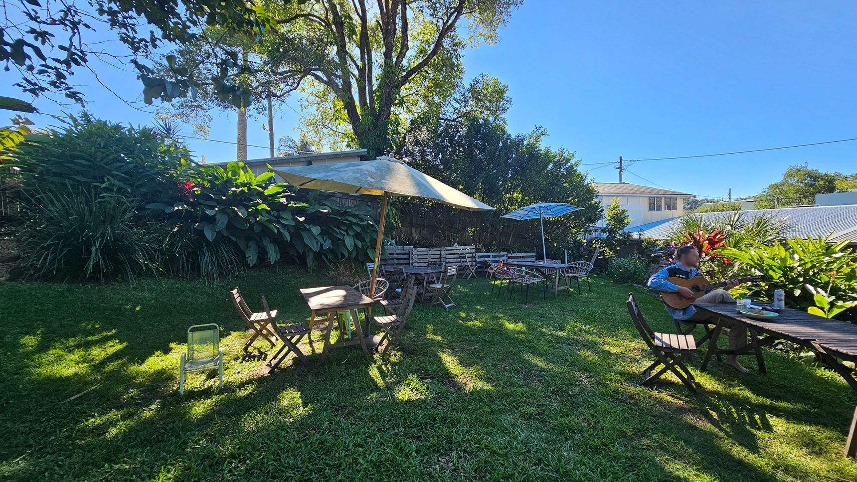 A garden setting with lush greenery and large plants in the background. Several wooden tables and chairs are scattered across the grass, with some tables shaded by large umbrellas. On the right, a person is seated at a table playing a guitar. A house with a sloped roof is visible in the background, partially obscured by foliage. The scene is well-lit, suggesting a sunny day. A garden setting with lush greenery and large plants in the background. Several wooden tables and chairs are scattered across the grass, with some tables shaded by large umbrellas. On the right, a person is seated at a table playing a guitar. A house with a sloped roof is visible in the background, partially obscured by foliage. The scene is well-lit, suggesting a sunny day.