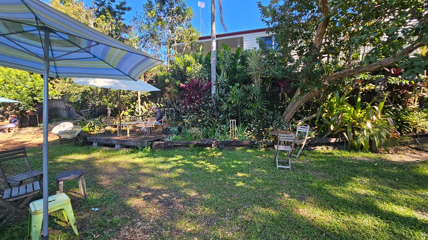 A garden setting with a grassy area in the foreground. On the left, a large striped umbrella shades a wooden chair and a small stool. In the centre, a wooden deck with a table and chairs is partially shaded by another umbrella. The background is filled with lush greenery, including various plants and trees. A house is partially visible behind the foliage. A few more chairs are scattered around the garden. A garden setting with a grassy area in the foreground. On the left, a large striped umbrella shades a wooden chair and a small stool. In the centre, a wooden deck with a table and chairs is partially shaded by another umbrella. The background is filled with lush greenery, including various plants and trees. A house is partially visible behind the foliage. A few more chairs are scattered around the garden.