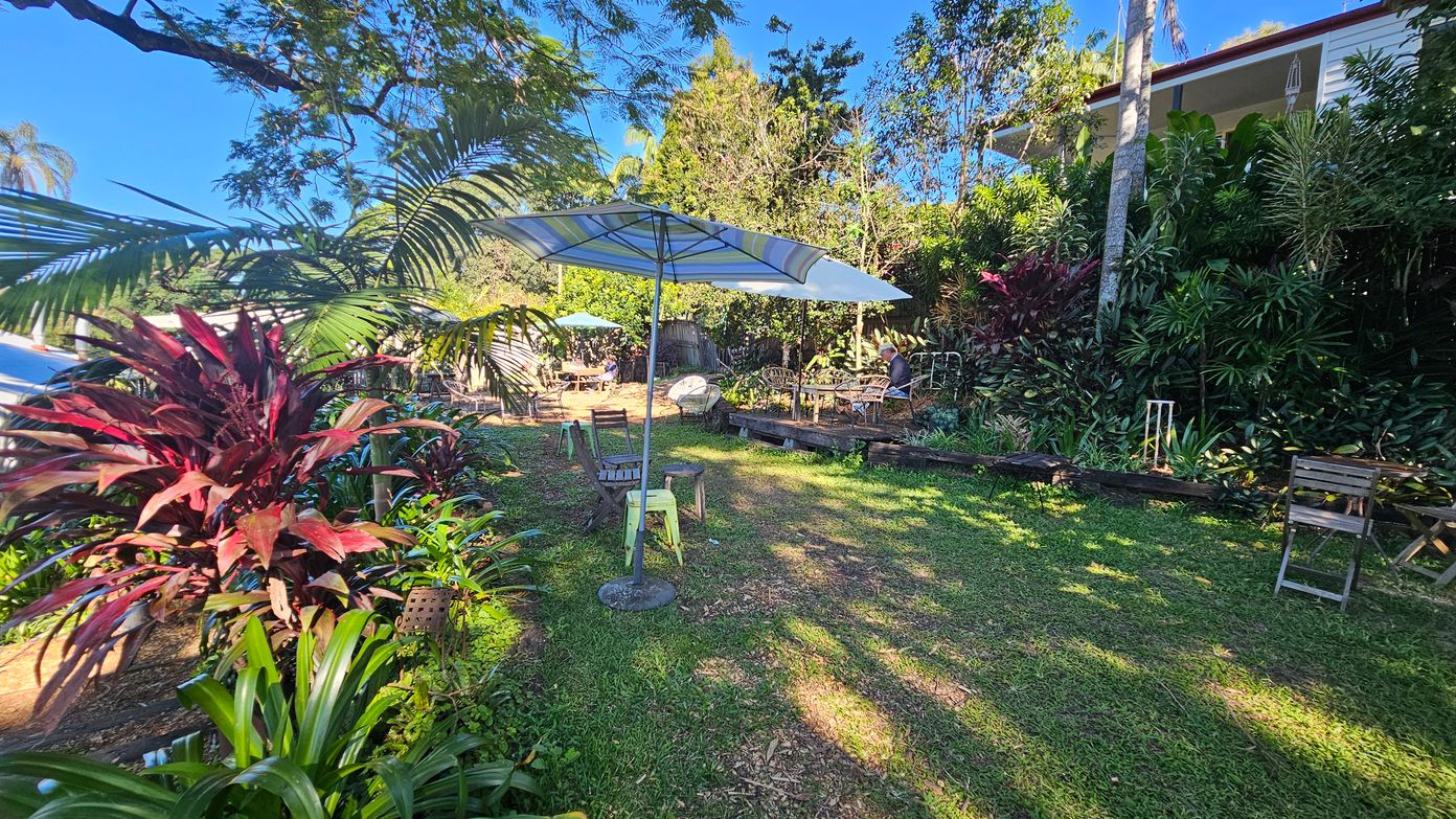 A garden with lush greenery and various plants, including red and green foliage on the left. In the centre, there are several outdoor tables and chairs, some with umbrellas providing shade. The ground is covered with grass, and there are patches of sunlight and shadow. In the background, a house is partially visible, surrounded by tall trees and dense vegetation. A garden with lush greenery and various plants, including red and green foliage on the left. In the centre, there are several outdoor tables and chairs, some with umbrellas providing shade. The ground is covered with grass, and there are patches of sunlight and shadow. In the background, a house is partially visible, surrounded by tall trees and dense vegetation.