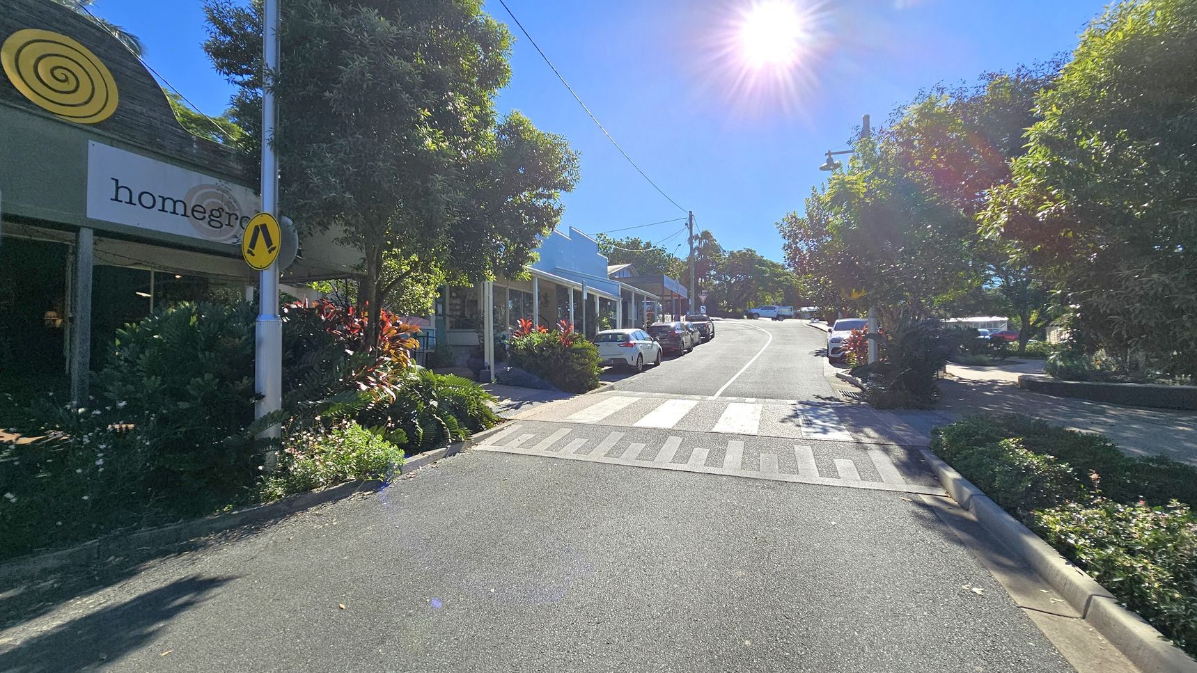 A street scene with a pedestrian crossing in the foreground. On the left, a building with a green exterior and a sign reading "homegrown" is partially visible, surrounded by lush greenery. On the right, a row of parked cars lines the street, which curves uphill. Trees and bushes are abundant on both sides, and the sun is shining brightly in the clear blue sky, casting shadows on the road. A street scene with a pedestrian crossing in the foreground. On the left, a building with a green exterior and a sign reading "homegrown" is partially visible, surrounded by lush greenery. On the right, a row of parked cars lines the street, which curves uphill. Trees and bushes are abundant on both sides, and the sun is shining brightly in the clear blue sky, casting shadows on the road.