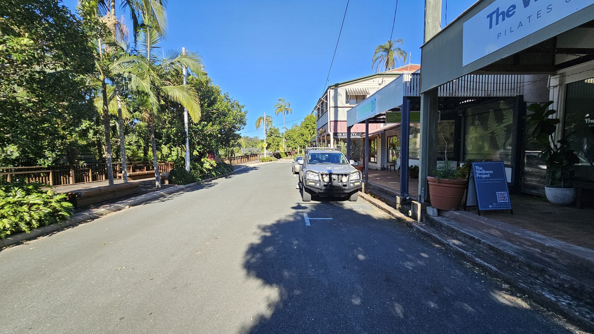 A street scene with a vehicle parked on the right side near a row of shops. The shop signs include "The Wellness Project" and "HB Beauty." Large potted plants are placed outside the shops. On the left, there are tall palm trees and dense greenery along a wooden fence. The road is clear, with shadows from the trees cast on the pavement. The sky is clear and blue. A street scene with a vehicle parked on the right side near a row of shops. The shop signs include "The Wellness Project" and "HB Beauty." Large potted plants are placed outside the shops. On the left, there are tall palm trees and dense greenery along a wooden fence. The road is clear, with shadows from the trees cast on the pavement. The sky is clear and blue.