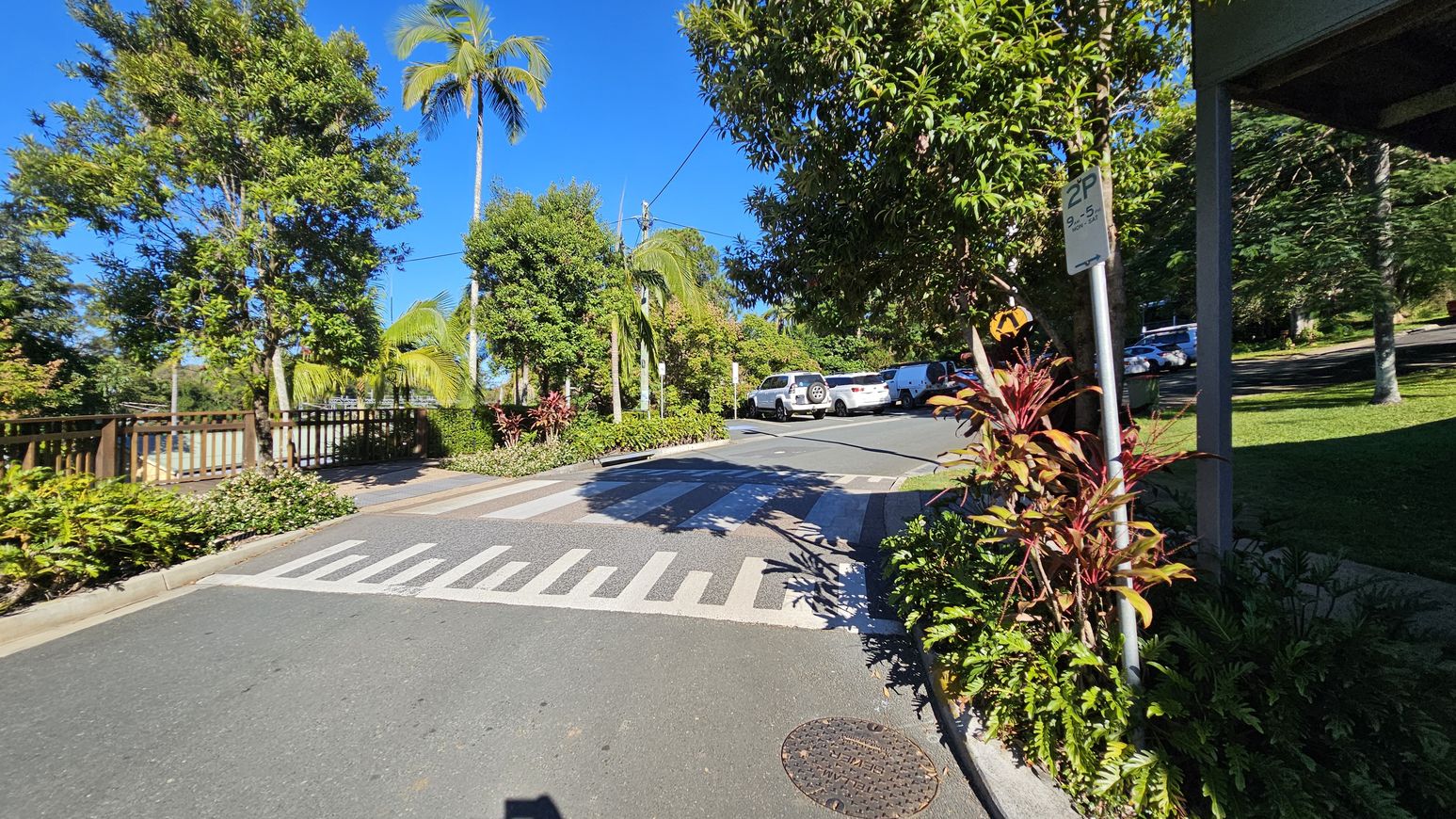 A street scene with a pedestrian crossing in the foreground. On the right, a signpost with parking information is surrounded by red and green foliage. Several cars are parked in a lot further back. On the left, a wooden fence runs alongside the road, with tall palm trees and lush greenery. The road surface is smooth, and the sky is clear and blue. A street scene with a pedestrian crossing in the foreground. On the right, a signpost with parking information is surrounded by red and green foliage. Several cars are parked in a lot further back. On the left, a wooden fence runs alongside the road, with tall palm trees and lush greenery. The road surface is smooth, and the sky is clear and blue.