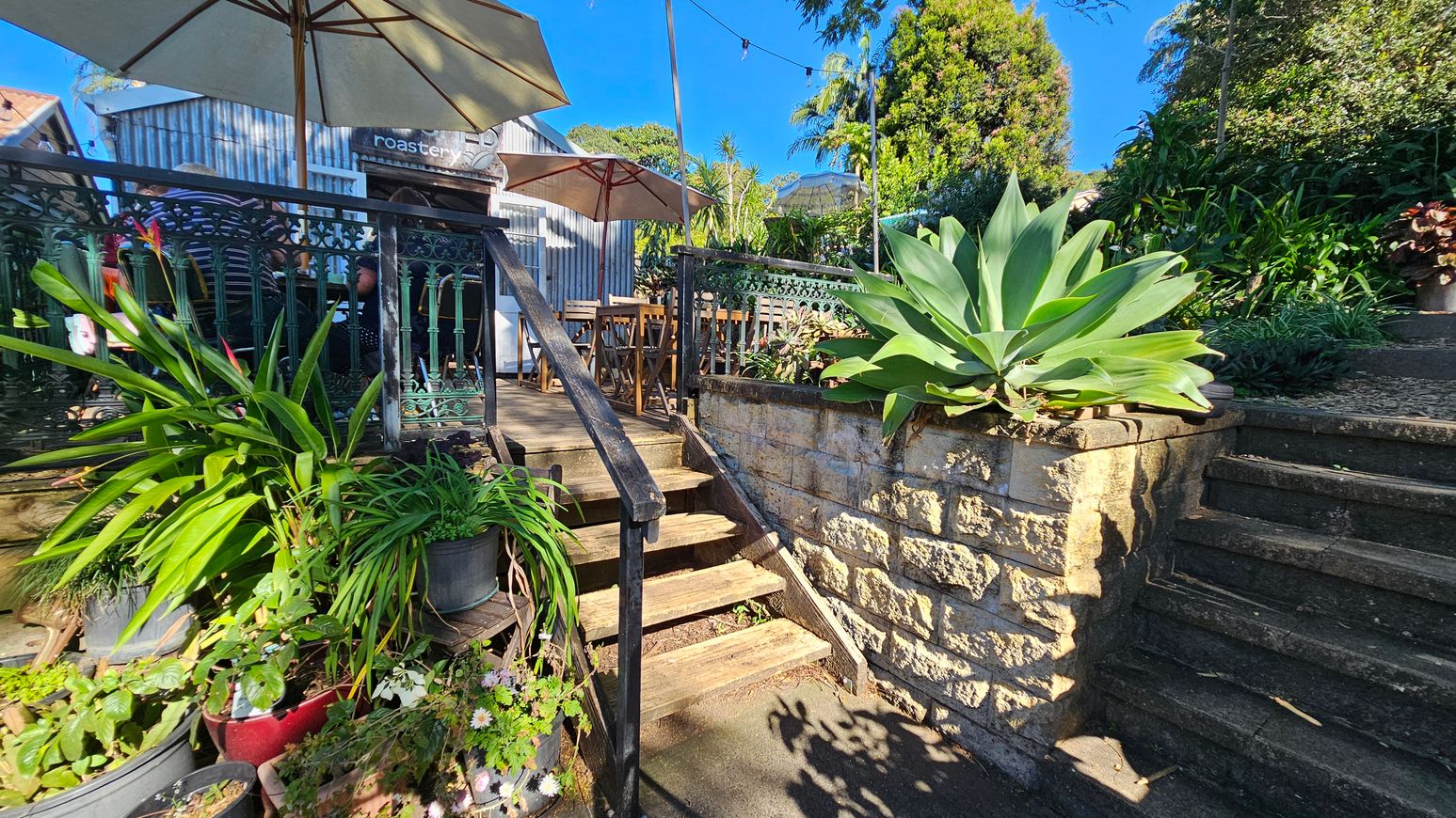 A small outdoor café area with wooden steps leading up to a deck on the left. The deck is bordered by a decorative metal railing and shaded by large umbrellas. Several potted plants, including a large succulent, are placed around the area. To the right, stone steps ascend alongside a stone wall, surrounded by lush greenery. The background features a corrugated metal building with a sign partially visible. The ground is paved, and string lights are strung above. A small outdoor café area with wooden steps leading up to a deck on the left. The deck is bordered by a decorative metal railing and shaded by large umbrellas. Several potted plants, including a large succulent, are placed around the area. To the right, stone steps ascend alongside a stone wall, surrounded by lush greenery. The background features a corrugated metal building with a sign partially visible. The ground is paved, and string lights are strung above.