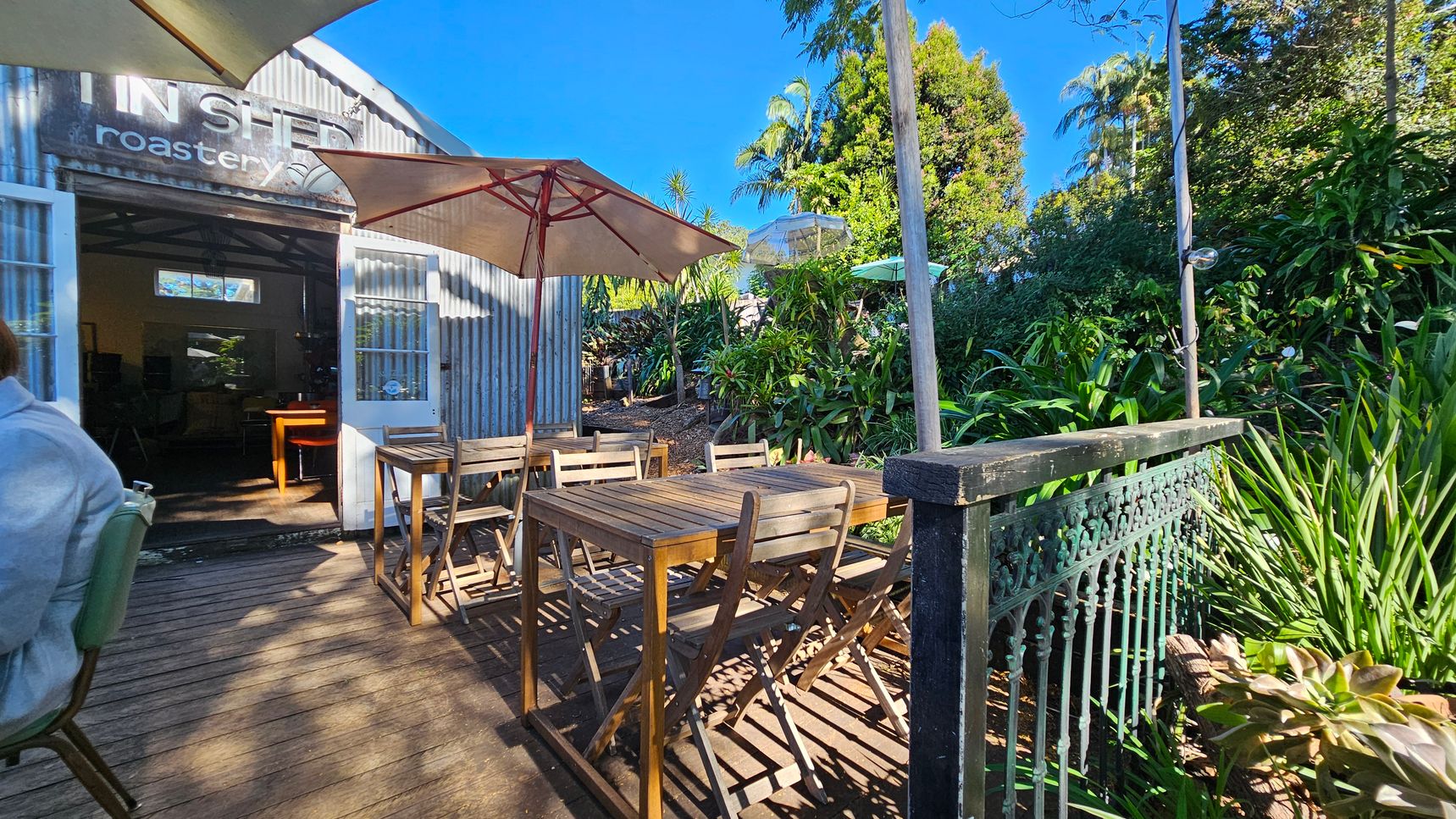 Outdoor seating area with wooden tables and chairs under large umbrellas. On the left, a corrugated metal building with an open door and a sign reading "roastery." Lush greenery and various plants surround the area, with a decorative metal railing on the right. The floor is wooden decking, and the sky is clear and blue. Outdoor seating area with wooden tables and chairs under large umbrellas. On the left, a corrugated metal building with an open door and a sign reading "roastery." Lush greenery and various plants surround the area, with a decorative metal railing on the right. The floor is wooden decking, and the sky is clear and blue.