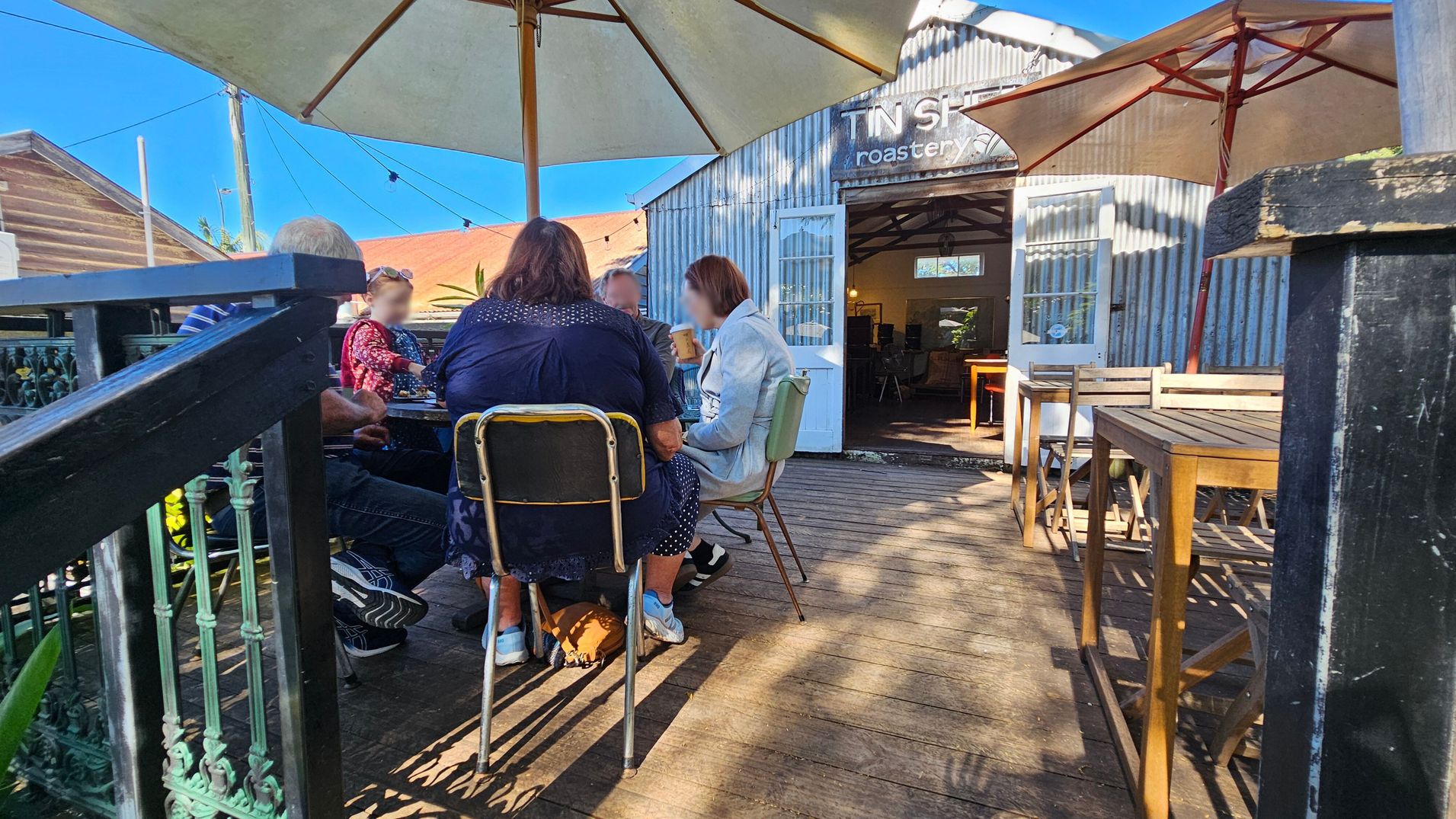 A group of people sit around a table under a large umbrella on a wooden deck. To the right, there are empty wooden tables and chairs. In the background, a corrugated metal building with an open door displays the words "Tin Shed Roastery." The deck is bordered by a decorative metal railing on the left. The scene is well-lit, suggesting a sunny day. A group of people sit around a table under a large umbrella on a wooden deck. To the right, there are empty wooden tables and chairs. In the background, a corrugated metal building with an open door displays the words "Tin Shed Roastery." The deck is bordered by a decorative metal railing on the left. The scene is well-lit, suggesting a sunny day.