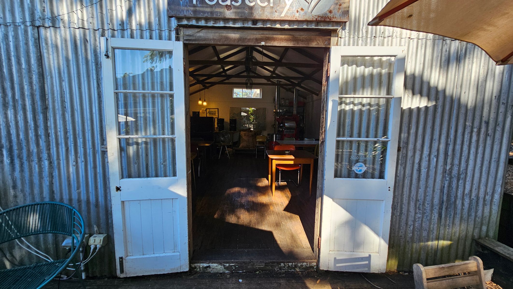 Corrugated metal building with open double doors leading inside. The doors are white with glass panes. Inside, a wooden table and chairs are visible on a wooden floor. The interior has a pitched roof with exposed beams. On the left, a blue metal bench is outside, and on the right, part of a wooden bench and an umbrella are visible. A sign above the doors reads "Roastery." Corrugated metal building with open double doors leading inside. The doors are white with glass panes. Inside, a wooden table and chairs are visible on a wooden floor. The interior has a pitched roof with exposed beams. On the left, a blue metal bench is outside, and on the right, part of a wooden bench and an umbrella are visible. A sign above the doors reads "Roastery."