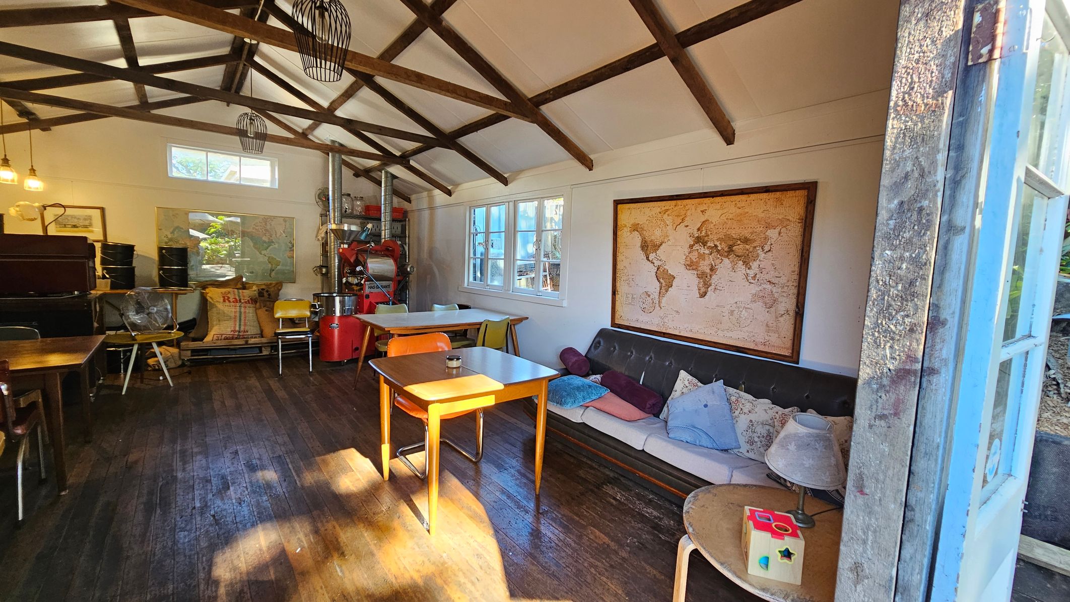 A rustic interior with wooden beams on the ceiling and a wooden floor. On the right, a sofa with assorted cushions is placed against the wall, above which hangs a large world map. A small table with a lamp and a toy is nearby. In the centre, a wooden table with chairs is illuminated by sunlight. On the left, there are more tables, chairs, and a coffee roaster. The walls have windows and framed pictures, and the space is lit by hanging lights. A rustic interior with wooden beams on the ceiling and a wooden floor. On the right, a sofa with assorted cushions is placed against the wall, above which hangs a large world map. A small table with a lamp and a toy is nearby. In the centre, a wooden table with chairs is illuminated by sunlight. On the left, there are more tables, chairs, and a coffee roaster. The walls have windows and framed pictures, and the space is lit by hanging lights.