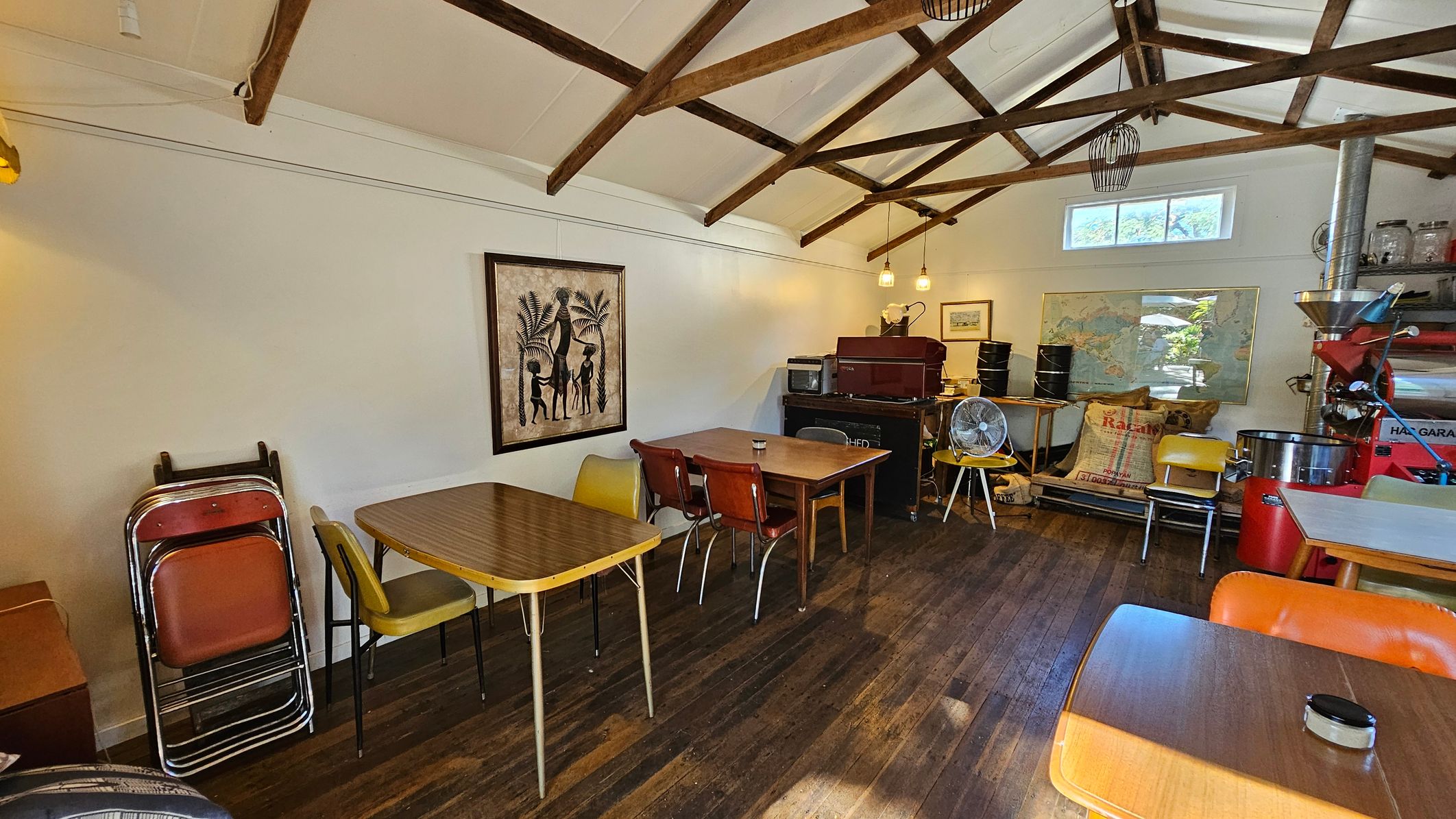 A cosy room with a wooden floor and exposed wooden beam ceiling. On the left, there are stacked chairs and a table with mismatched chairs. A framed artwork depicting figures and palm trees hangs on the wall. In the centre, more tables and chairs are arranged, with a small fan on one table. The right side features a coffee roaster, bags of coffee beans, and a map on the wall. Pendant lights hang from the ceiling, and a small window lets in natural light. A cosy room with a wooden floor and exposed wooden beam ceiling. On the left, there are stacked chairs and a table with mismatched chairs. A framed artwork depicting figures and palm trees hangs on the wall. In the centre, more tables and chairs are arranged, with a small fan on one table. The right side features a coffee roaster, bags of coffee beans, and a map on the wall. Pendant lights hang from the ceiling, and a small window lets in natural light.