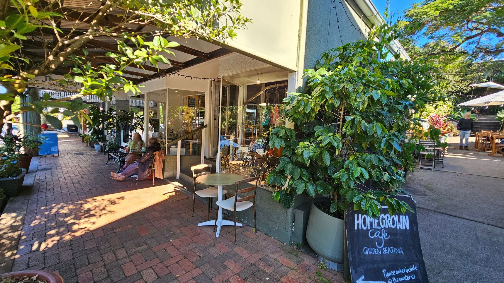 A café with outdoor seating is on the right, featuring a chalkboard sign that reads "Homegrown Café Garden Seating." Potted plants and a small table with chairs are nearby. On the left, a brick pathway is lined with more plants and a few people sitting on benches. The café has large windows, and the area is shaded by a wooden pergola. In the background, there are more tables with umbrellas and a person walking away. A café with outdoor seating is on the right, featuring a chalkboard sign that reads "Homegrown Café Garden Seating." Potted plants and a small table with chairs are nearby. On the left, a brick pathway is lined with more plants and a few people sitting on benches. The café has large windows, and the area is shaded by a wooden pergola. In the background, there are more tables with umbrellas and a person walking away.