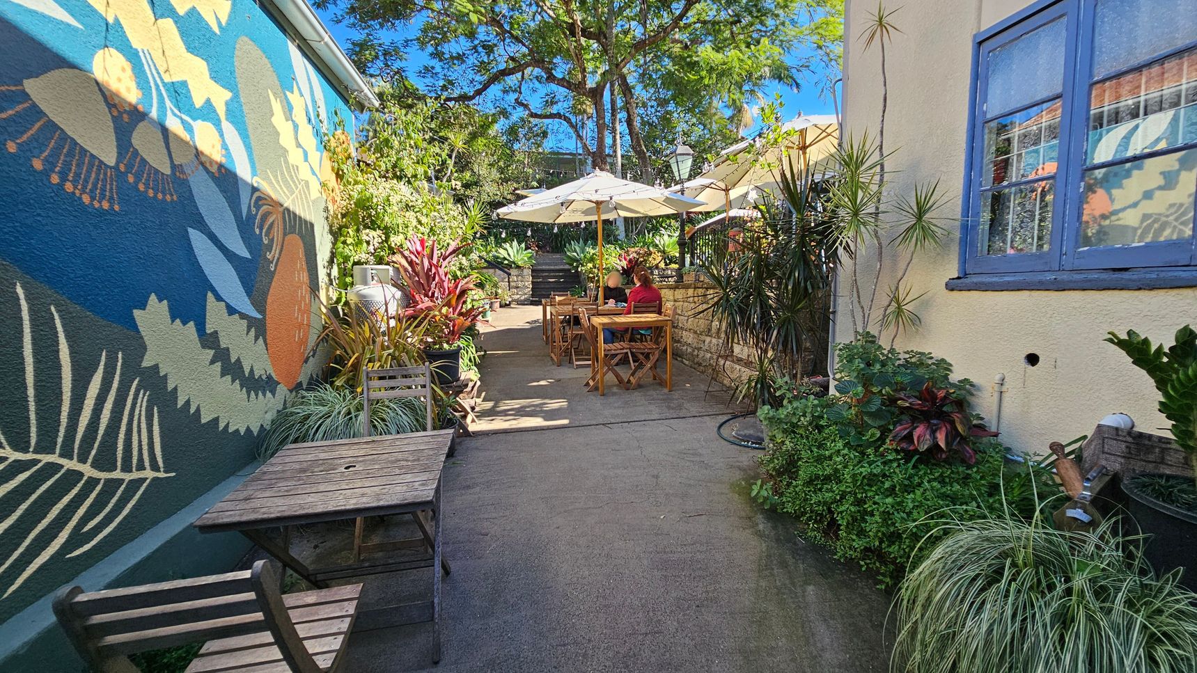 A narrow outdoor café area with a colourful mural on the left wall featuring abstract plant designs. Wooden tables and chairs are placed along the path, with a couple seated at one table under large white umbrellas. Lush greenery and potted plants line both sides, creating a vibrant atmosphere. The right side has a light-coloured wall with a blue-framed window. In the background, a tree provides shade, and steps lead to another area. The ground is paved with concrete. A narrow outdoor café area with a colourful mural on the left wall featuring abstract plant designs. Wooden tables and chairs are placed along the path, with a couple seated at one table under large white umbrellas. Lush greenery and potted plants line both sides, creating a vibrant atmosphere. The right side has a light-coloured wall with a blue-framed window. In the background, a tree provides shade, and steps lead to another area. The ground is paved with concrete.