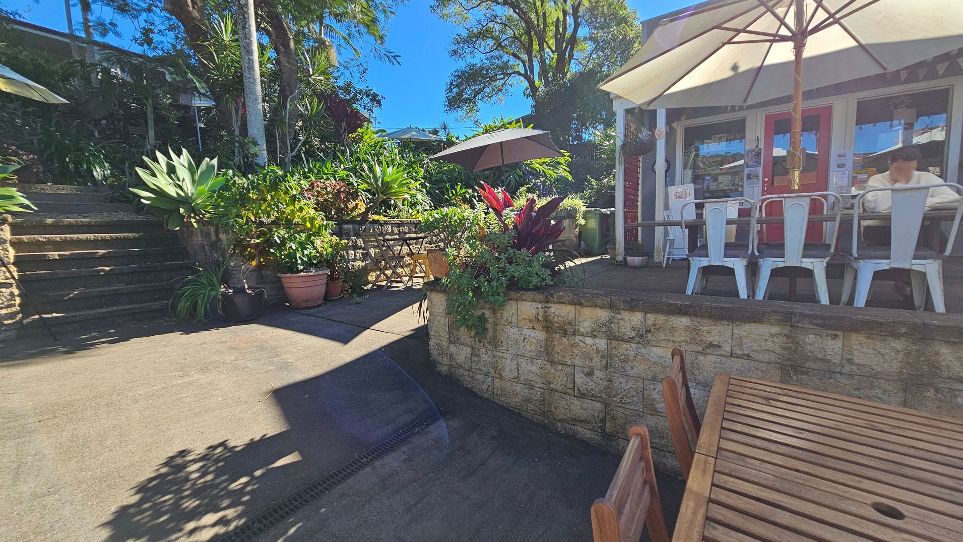 Outdoor café setting with a wooden table and chairs in the foreground. On the right, a café with a red door and white-framed windows, featuring metal chairs and a large umbrella. A person is seated at a table. In the middle, a stone wall with lush green plants and red foliage. On the left, stone steps lead up to more greenery and a shaded area with additional tables and chairs. Tall trees and a clear blue sky are visible in the background. Outdoor café setting with a wooden table and chairs in the foreground. On the right, a café with a red door and white-framed windows, featuring metal chairs and a large umbrella. A person is seated at a table. In the middle, a stone wall with lush green plants and red foliage. On the left, stone steps lead up to more greenery and a shaded area with additional tables and chairs. Tall trees and a clear blue sky are visible in the background.