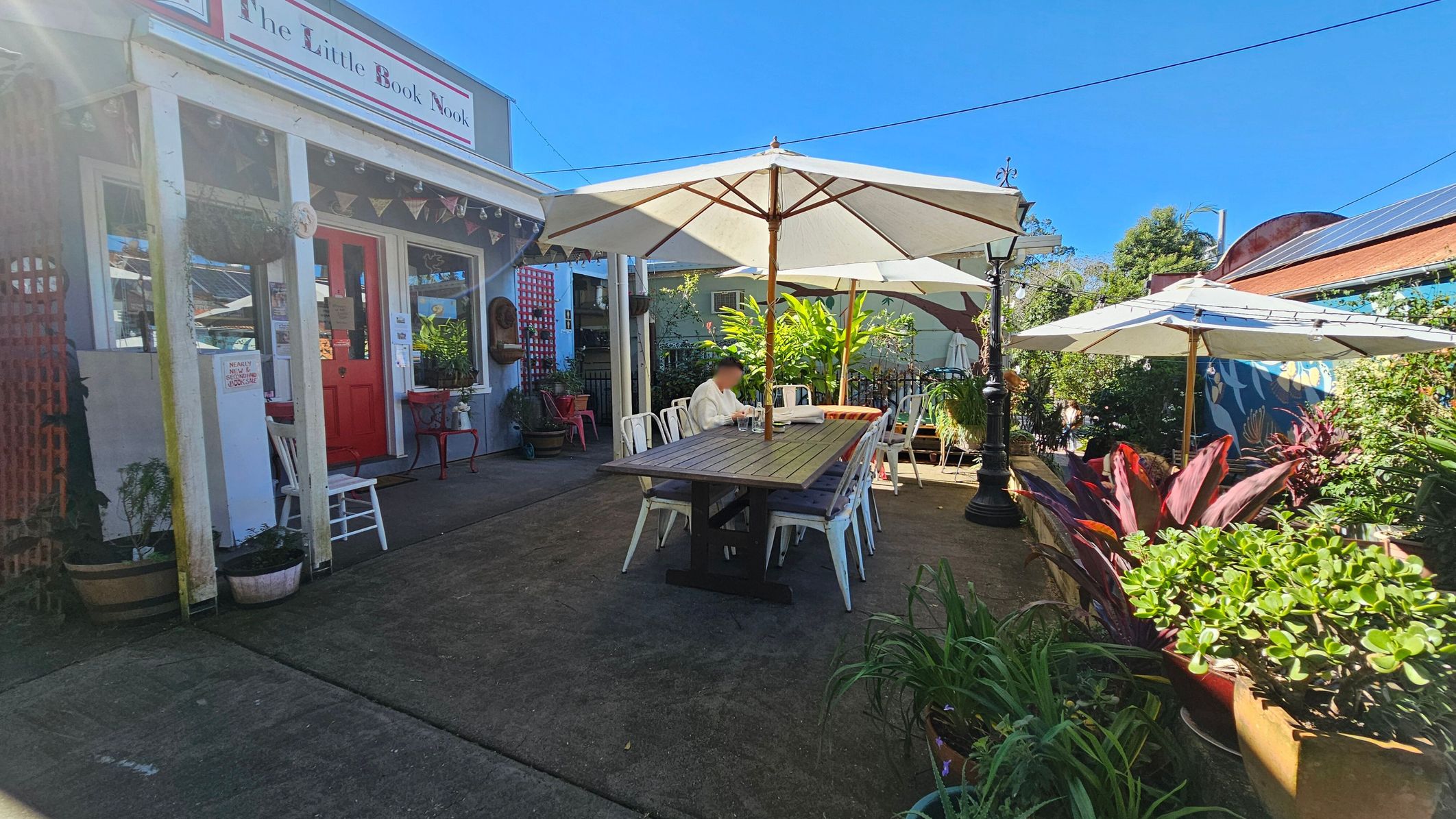 A quaint outdoor seating area with a long wooden table and white chairs under large white umbrellas. On the left, a building with a red door and a sign reading "The Little Book Nook" is adorned with bunting. Potted plants and greenery surround the area, adding a lush feel. A person is seated at the table, and a lamppost stands nearby. The ground is paved, and the background features a mural and more plants. A quaint outdoor seating area with a long wooden table and white chairs under large white umbrellas. On the left, a building with a red door and a sign reading "The Little Book Nook" is adorned with bunting. Potted plants and greenery surround the area, adding a lush feel. A person is seated at the table, and a lamppost stands nearby. The ground is paved, and the background features a mural and more plants.