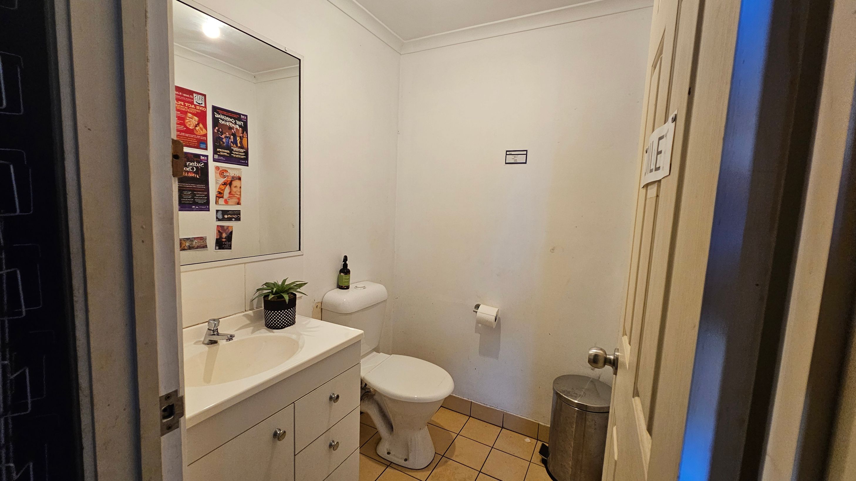 A small bathroom with white walls and a tiled floor. On the left, a white vanity with a sink, a small plant, and a mirror above. The toilet is positioned in the centre against the back wall, with a toilet roll holder to the right. A silver bin is on the floor near the toilet. Posters are visible on the wall reflected in the mirror. The door on the right has a sign with the word "TOILET". The ceiling is plain with a light fixture. A small bathroom with white walls and a tiled floor. On the left, a white vanity with a sink, a small plant, and a mirror above. The toilet is positioned in the centre against the back wall, with a toilet roll holder to the right. A silver bin is on the floor near the toilet. Posters are visible on the wall reflected in the mirror. The door on the right has a sign with the word "TOILET". The ceiling is plain with a light fixture.
