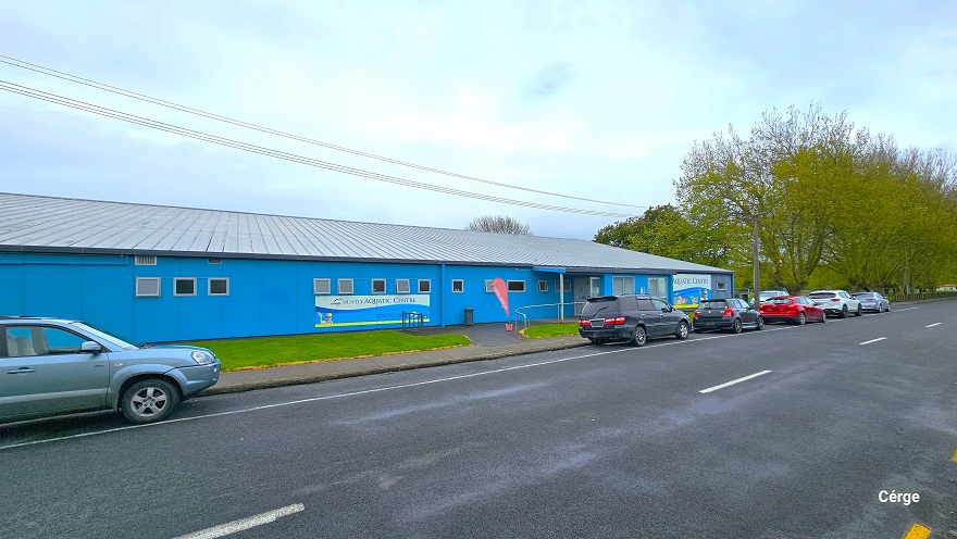 View of the parking area of the Aquatic Centre along the road of Wight Street, where several cars are parked. The low-set blue building can be seen in the background, with numerous trees located in the far right corner. There is a small white signage on the front blue wall detailing the name of the facility and a red, teardrop-shaped flag at the entryway to the facility. View of the parking area of the Aquatic Centre along the road of Wight Street, where several cars are parked. The low-set blue building can be seen in the background, with numerous trees located in the far right corner. There is a small white signage on the front blue wall detailing the name of the facility and a red, teardrop-shaped flag at the entryway to the facility.