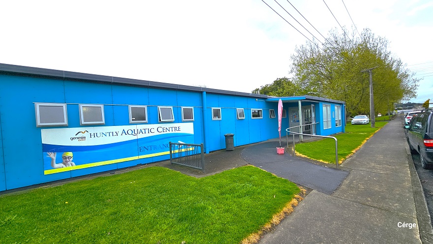 A closer look at the entrance leading to the Huntly Aquatic Centre. A dark grey pathway with steel handrails connects the sidewalk to the main glass entrance doors of the center. Small square glass windows are placed alongside the front wall of the center. Cars are parked in front, and to the back is a white car. The landscape of the area is well maintained as grasses are at the same height and can be distinguished by a yellow marking on the side. A closer look at the entrance leading to the Huntly Aquatic Centre. A dark grey pathway with steel handrails connects the sidewalk to the main glass entrance doors of the center. Small square glass windows are placed alongside the front wall of the center. Cars are parked in front, and to the back is a white car. The landscape of the area is well maintained as grasses are at the same height and can be distinguished by a yellow marking on the side.