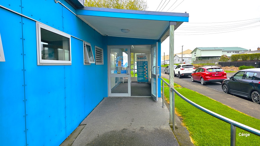 View of the entrance glass doors at Huntly Aquatic Centre coming from the cemented pathway with steel handrails. The main entrance has two glass doors lined with white painted panels, with the right door open. The small glass windows on the blue-painted wall to the left are slightly open. Grey, black, red, and white cars are parked in front. Residential buildings are noticeable in the far right corner of the image. View of the entrance glass doors at Huntly Aquatic Centre coming from the cemented pathway with steel handrails. The main entrance has two glass doors lined with white painted panels, with the right door open. The small glass windows on the blue-painted wall to the left are slightly open. Grey, black, red, and white cars are parked in front. Residential buildings are noticeable in the far right corner of the image.