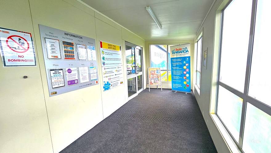 An inside look at the space after passing through the main entrance doors of Huntly Aquatic Centre. The floor is covered in a grey mat with a parallel line design. On the left wall are white, small signs for “No Bombing”, the information bulletin, and the Huntly Pool Safety Rules. On the right are tempered glass windows for privacy inside. Two advertising sign boards are placed in front of another entry door inside the centre. An inside look at the space after passing through the main entrance doors of Huntly Aquatic Centre. The floor is covered in a grey mat with a parallel line design. On the left wall are white, small signs for “No Bombing”, the information bulletin, and the Huntly Pool Safety Rules. On the right are tempered glass windows for privacy inside. Two advertising sign boards are placed in front of another entry door inside the centre.