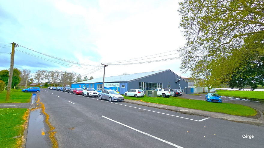 Opposite view of the entire front and side of the Huntly Aquatic Centre as well as the entire Wight Street. The front wall is painted in blue, while the right wall is painted in grey. The entire building has multiple glass windows. The grey wall has tall rectangular glass windows, while the front blue wall has a mixture of small and tall glass windows. Numerous vehicles are parked in front, each facing different directions. Power poles line both sides of the street. Opposite view of the entire front and side of the Huntly Aquatic Centre as well as the entire Wight Street. The front wall is painted in blue, while the right wall is painted in grey. The entire building has multiple glass windows. The grey wall has tall rectangular glass windows, while the front blue wall has a mixture of small and tall glass windows. Numerous vehicles are parked in front, each facing different directions. Power poles line both sides of the street.