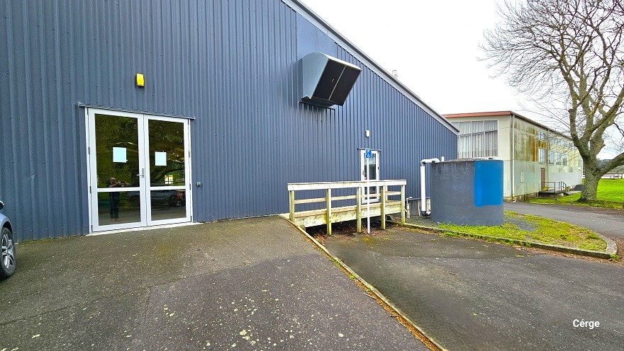 A closer look at the side entry glass doors leading inside the Huntly Aquatic Centre. The walls are painted in a dark purple hue. The inclined ramp leading to the glass doors is cemented. To the right is another glass door with white linings on the sides and white railings. There is a curvy elevation on the upper part of the wall for exhaust. Further to the right is a large circular tank with white PVC tubes, and to the back is a separate building. A closer look at the side entry glass doors leading inside the Huntly Aquatic Centre. The walls are painted in a dark purple hue. The inclined ramp leading to the glass doors is cemented. To the right is another glass door with white linings on the sides and white railings. There is a curvy elevation on the upper part of the wall for exhaust. Further to the right is a large circular tank with white PVC tubes, and to the back is a separate building.