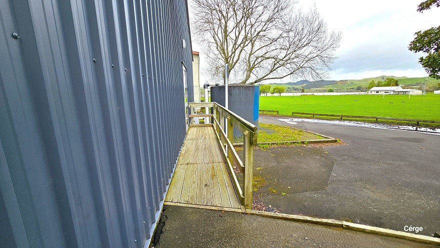 A closer look at the narrow space within the white railings of the side of Huntly Aquatic Centre. The floor space is made of wood as well as the handrails connected to the inclined cemented ramp. From this position, a large, open space of green field enclosed by small wooden fences can be seen. Towards the back is a huge tank with white PVC tubes, a large dead tree, and a glimpse of part of a white building to the back. A closer look at the narrow space within the white railings of the side of Huntly Aquatic Centre. The floor space is made of wood as well as the handrails connected to the inclined cemented ramp. From this position, a large, open space of green field enclosed by small wooden fences can be seen. Towards the back is a huge tank with white PVC tubes, a large dead tree, and a glimpse of part of a white building to the back.