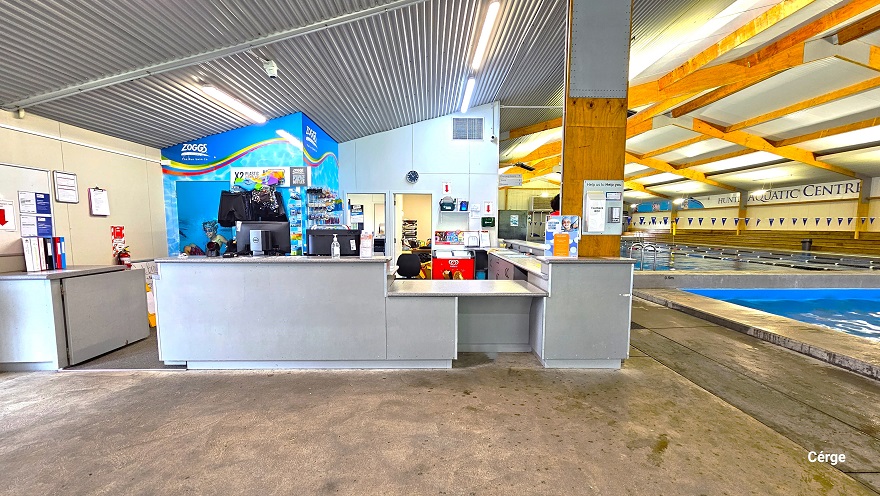 The image shows the double entrance glass doors, the reception desk, and the two pools. On the side of the reception desk are two young lifeguard males wearing orange and yellow uniforms. There is an adequate space that allows access surrounding each pool. Opposite to the two lifeguards is a pool with a 0.9-meter depth. The roof structure is made of corrugated iron and is well-lit with fluorescent lighting. The image shows the double entrance glass doors, the reception desk, and the two pools. On the side of the reception desk are two young lifeguard males wearing orange and yellow uniforms. There is an adequate space that allows access surrounding each pool. Opposite to the two lifeguards is a pool with a 0.9-meter depth. The roof structure is made of corrugated iron and is well-lit with fluorescent lighting.