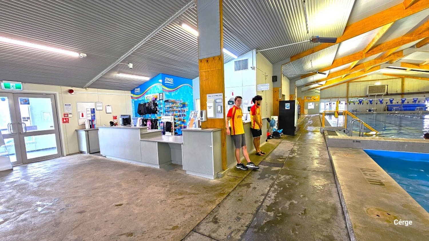 Front view of the white-to-grey reception desk area inside the Huntly Aquatic Centre. The reception area offers swimwear and other gear for sale. A water-like blue wall and a small office space at the back distinguish the reception area. On the left is a whiteboard with documents hanging next to a fire extinguisher. On the right are two pools with blue floating rope lanes, a string of blue triangular flags, and large, brown wooden frames that light the ceiling. Front view of the white-to-grey reception desk area inside the Huntly Aquatic Centre. The reception area offers swimwear and other gear for sale. A water-like blue wall and a small office space at the back distinguish the reception area. On the left is a whiteboard with documents hanging next to a fire extinguisher. On the right are two pools with blue floating rope lanes, a string of blue triangular flags, and large, brown wooden frames that light the ceiling.
