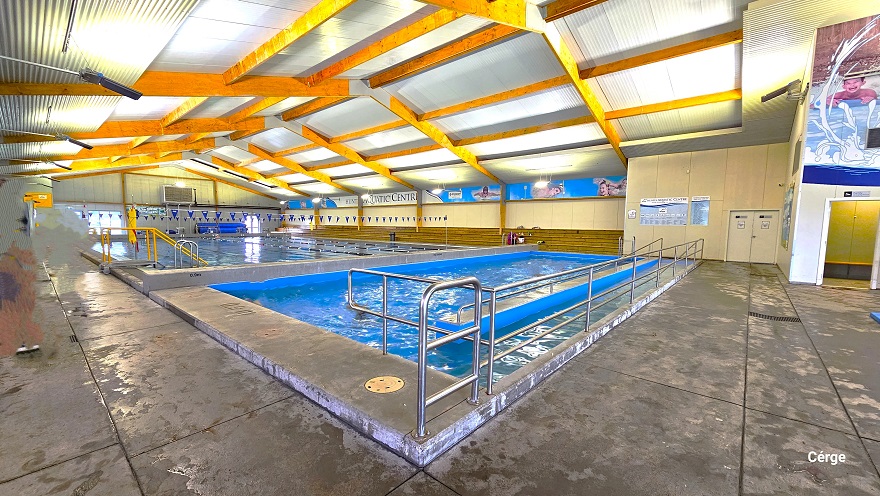 A closer view of the ramp access with steel handrails of another small pool in Huntly Aquatic Centre. The ceiling has brown wooden support beams that support the white corrugated roof. Next to this small pool is a large pool with blue and white floating rope lanes. Four ladder accesses are marked by an inverted “U” steel frame on the pool sides. Long rows of low brown benches are placed near the large pool. A closer view of the ramp access with steel handrails of another small pool in Huntly Aquatic Centre. The ceiling has brown wooden support beams that support the white corrugated roof. Next to this small pool is a large pool with blue and white floating rope lanes. Four ladder accesses are marked by an inverted “U” steel frame on the pool sides. Long rows of low brown benches are placed near the large pool.