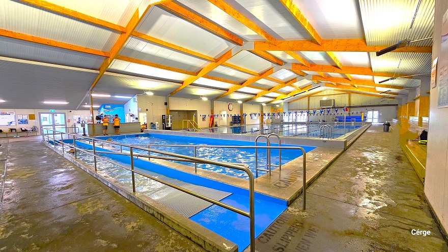Closer view of the ramp access with steel handrails. There is a grey mat along the ramp access with small circular bumps for added friction to the feet to support the guest while entering the pool. The pool has deep blue walls, and within the cemented space surrounding the pool is the word “Caution: Slippery when wet”. A black waste bin is visible in the far-right area of the pathway. Strings of triangular blue flags can be seen hanging above the large pool. Closer view of the ramp access with steel handrails. There is a grey mat along the ramp access with small circular bumps for added friction to the feet to support the guest while entering the pool. The pool has deep blue walls, and within the cemented space surrounding the pool is the word “Caution: Slippery when wet”. A black waste bin is visible in the far-right area of the pathway. Strings of triangular blue flags can be seen hanging above the large pool.