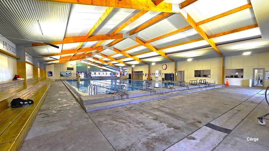 View of the large pool near the long row of low-set brown benches. On the upper wall of the back of these benches is a large font of Huntly Aquatic Centre. Opposite this wall is the wall with a large red circular clock with two open doors for bathrooms and changing rooms. Starting blocks are lined at the elevated edge. View of the large pool near the long row of low-set brown benches. On the upper wall of the back of these benches is a large font of Huntly Aquatic Centre. Opposite this wall is the wall with a large red circular clock with two open doors for bathrooms and changing rooms. Starting blocks are lined at the elevated edge.