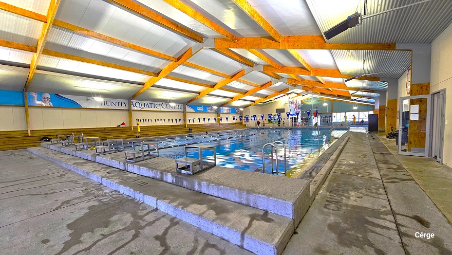 A closer look at the elevated edge at the starting point of the large pool. Grey diving blocks are placed within each pool lane. The white ceiling is corrugated designed with large wooden beams. Strings of triangular blue flags can be seen hanging above the large pool. On the right is an open glass door with small papers attached to it. A closer look at the elevated edge at the starting point of the large pool. Grey diving blocks are placed within each pool lane. The white ceiling is corrugated designed with large wooden beams. Strings of triangular blue flags can be seen hanging above the large pool. On the right is an open glass door with small papers attached to it.