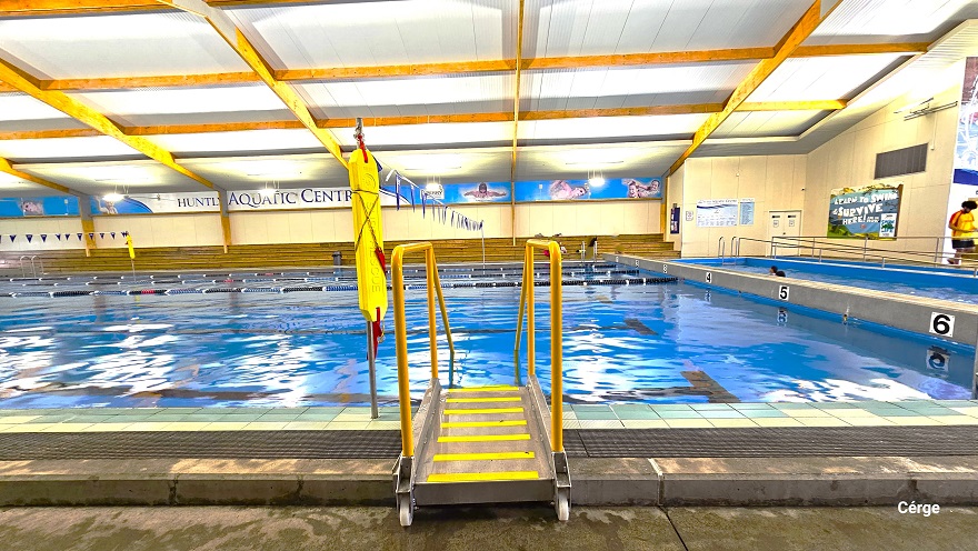 One of the access pool ladders with yellow hand railings on the side of the large pool at Huntly Aquatic Centre. On the pool wall, a few centimeters above the pool water, are numbers from 1-6 assigned for each pool lane. The wall further to the back, on the right side, is mostly covered by informative graphics. The name of the centre is clearly visible on the upper part of the wall directly opposite. One of the access pool ladders with yellow hand railings on the side of the large pool at Huntly Aquatic Centre. On the pool wall, a few centimeters above the pool water, are numbers from 1-6 assigned for each pool lane. The wall further to the back, on the right side, is mostly covered by informative graphics. The name of the centre is clearly visible on the upper part of the wall directly opposite.