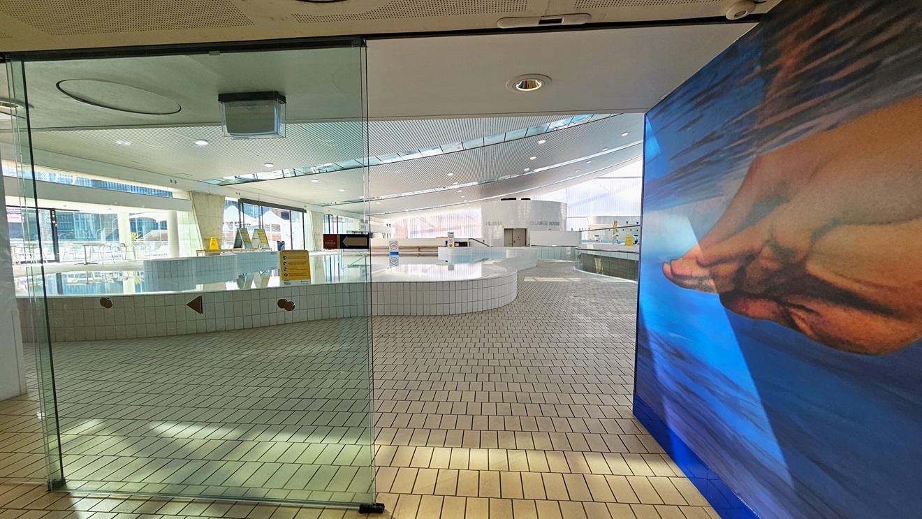 Access to the aquatic center through an automatic glass sliding door. The right wall features a life-sized painting of Ian Thorpe swimming underwater. Inside, the leisure pool with stairs and a handrail is directly ahead. The floor is tiled with small bricks, and the ceiling displays a white porous design with circular LED lights. Access to the aquatic center through an automatic glass sliding door. The right wall features a life-sized painting of Ian Thorpe swimming underwater. Inside, the leisure pool with stairs and a handrail is directly ahead. The floor is tiled with small bricks, and the ceiling displays a white porous design with circular LED lights.